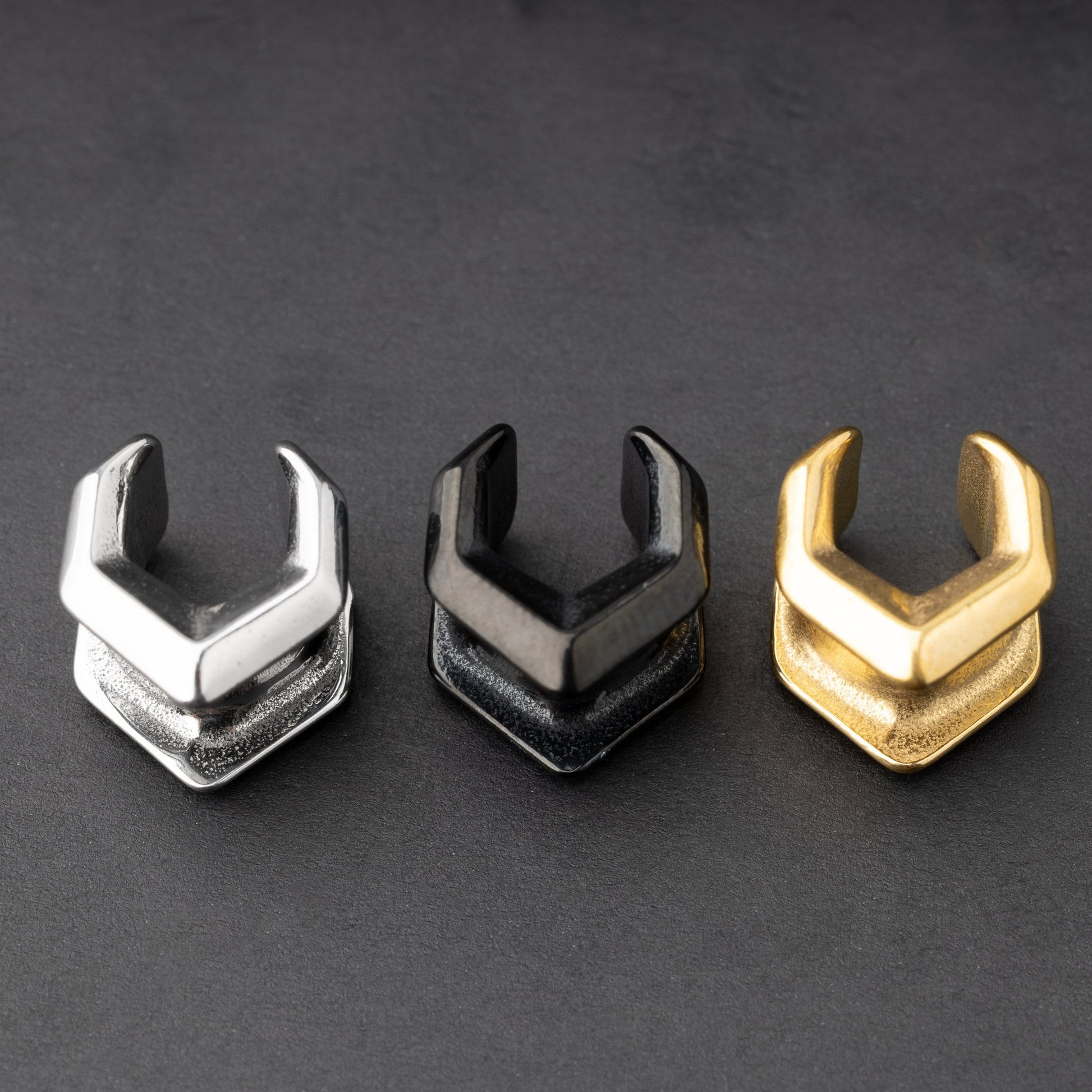 Three geometric chevron-shaped ear cuffs in silver, black, and gold finishes, displayed on a dark background.