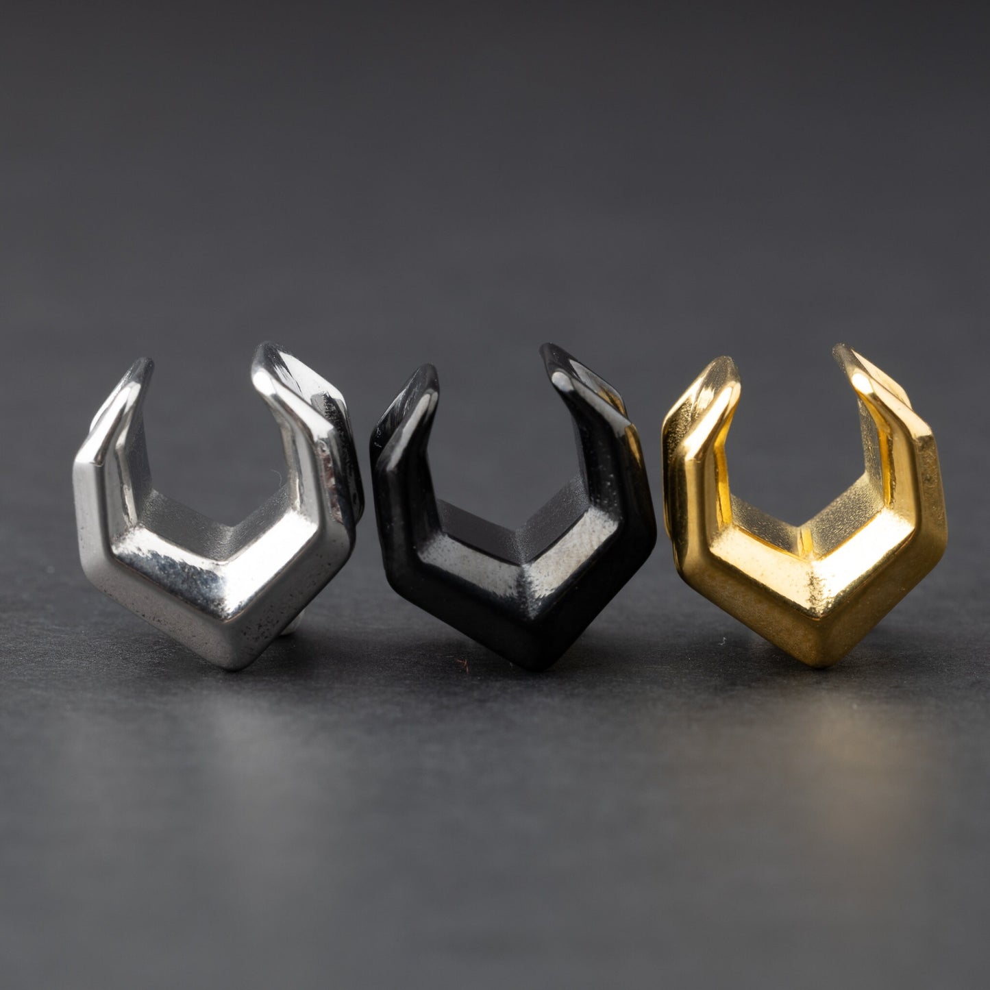 Three geometric chevron-shaped ear cuffs in silver, black, and gold finishes, displayed on a dark background.