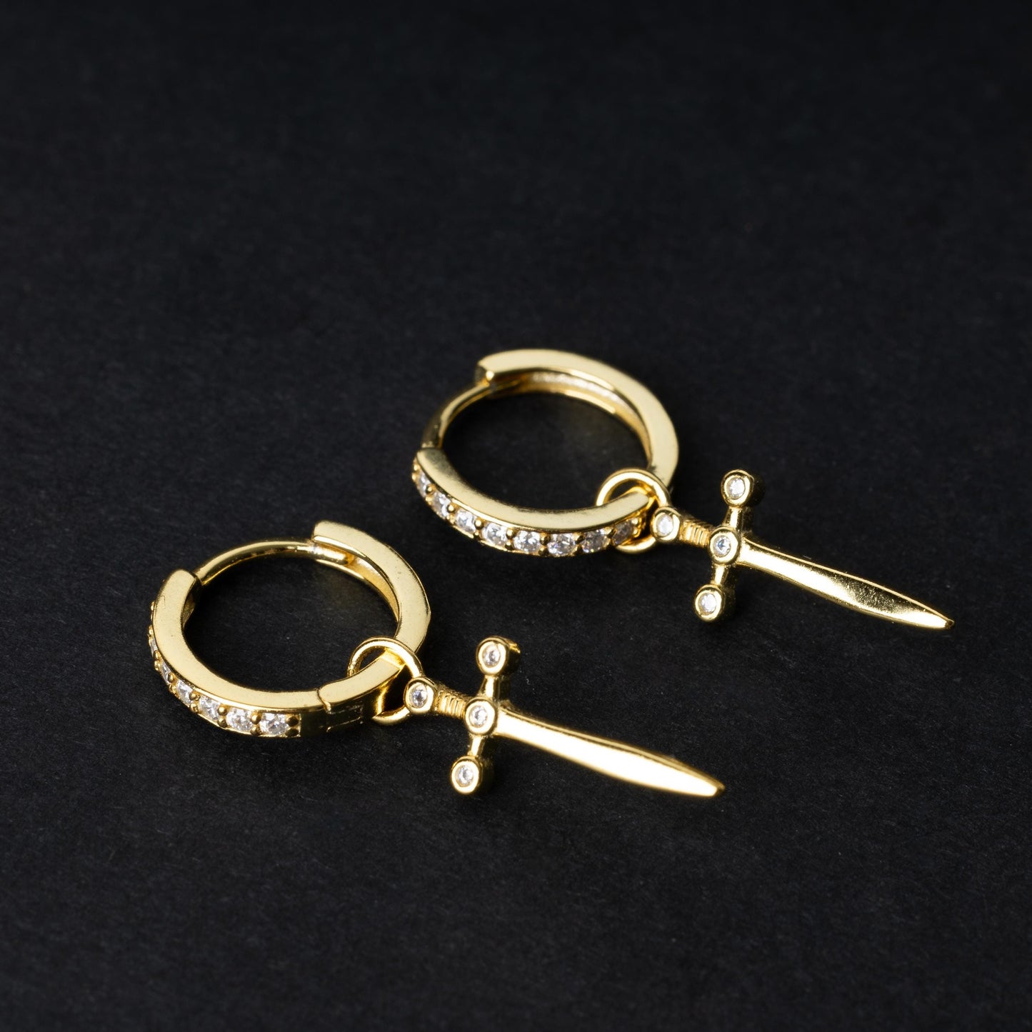 Gold-plated hoop earrings with crystal detailing and sword-shaped charms, displayed on a dark background.