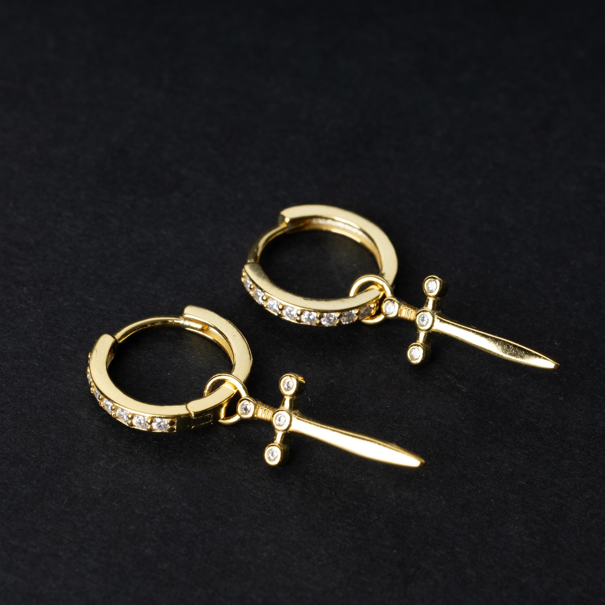 Gold-plated hoop earrings with crystal detailing and sword-shaped charms, displayed on a dark background.