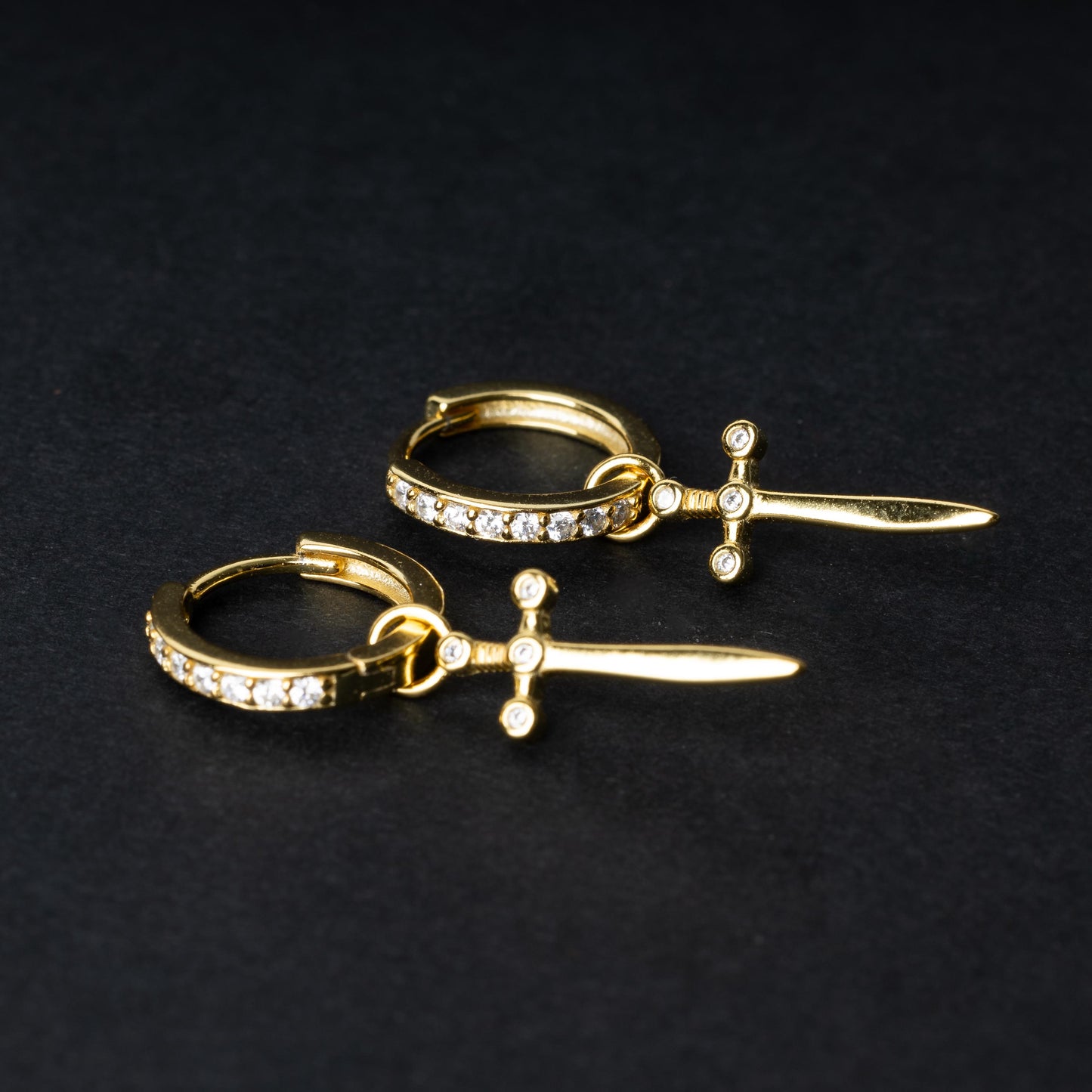 Gold-plated hoop earrings with crystal detailing and sword-shaped charms, displayed on a dark background.