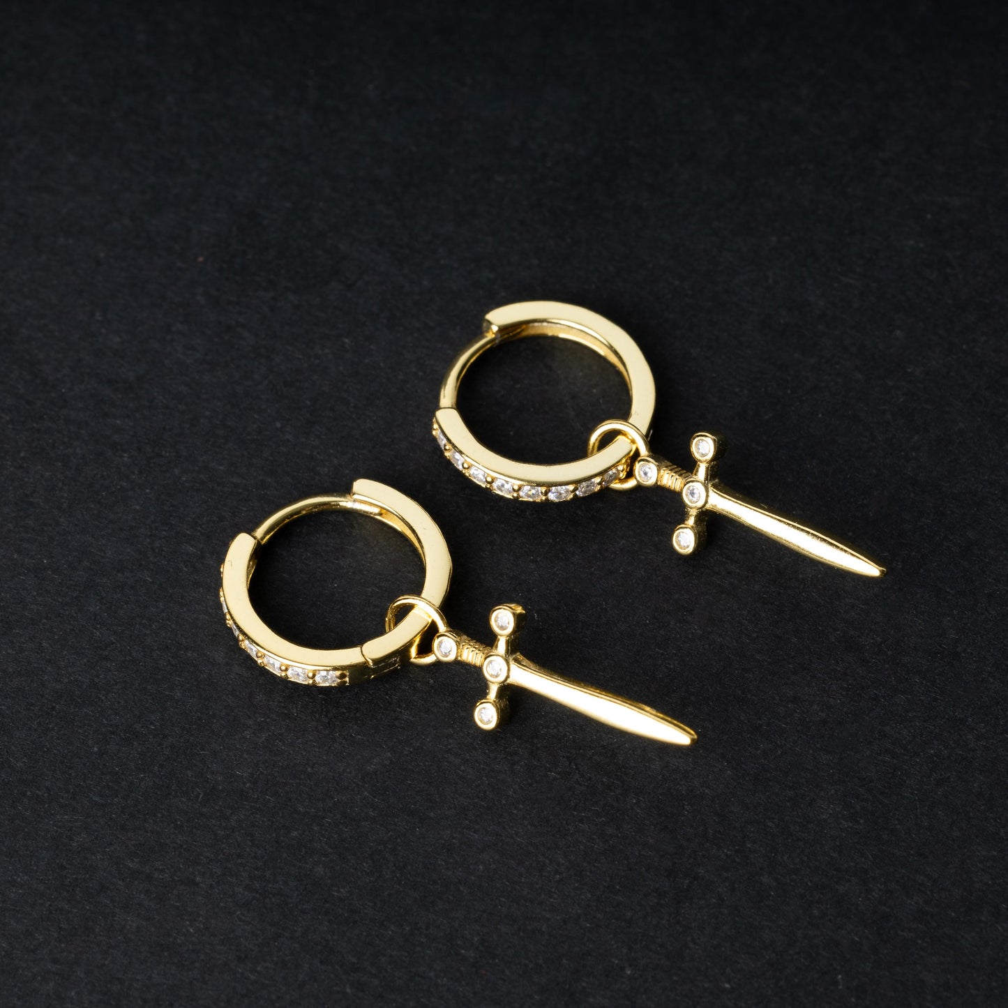 Gold-plated hoop earrings with crystal detailing and sword-shaped charms, displayed on a dark background.