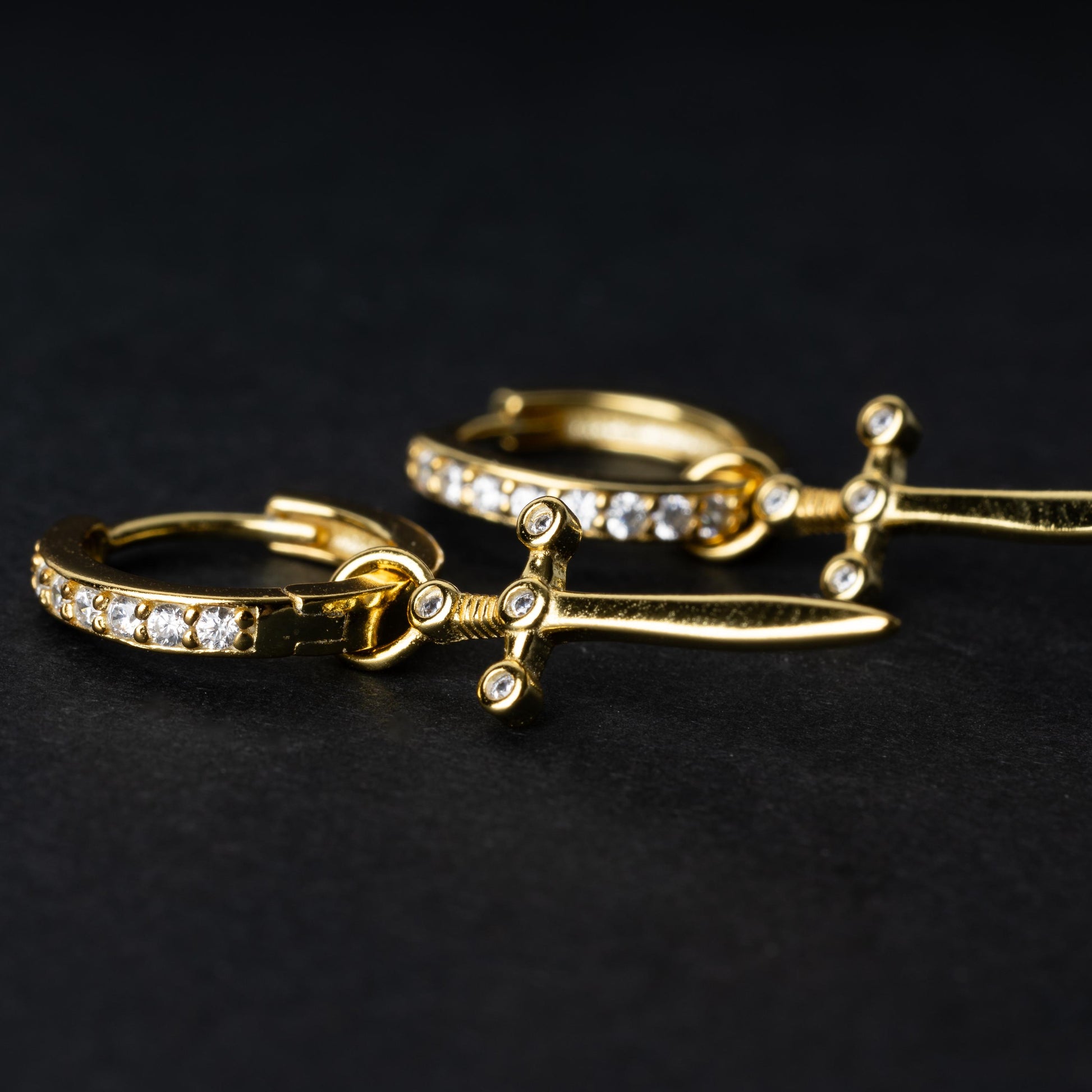 Close up of Gold-plated hoop earrings with crystal detailing and sword-shaped charms, displayed on a dark background.