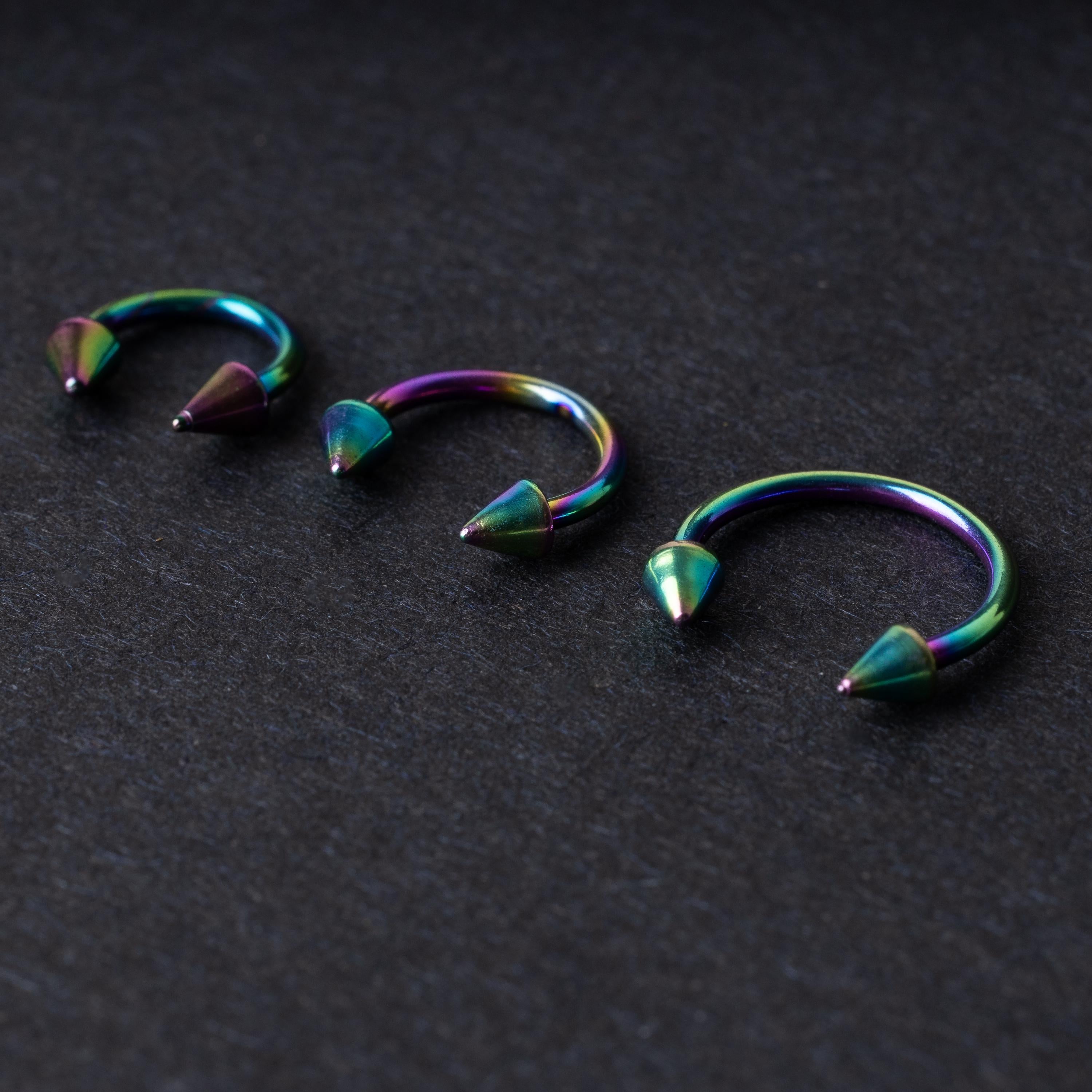 Three stainless steel horseshoe hoops with cone-shaped ends and a rainbow anodized finish displayed on a dark background, showcasing their bold design and vibrant colors.