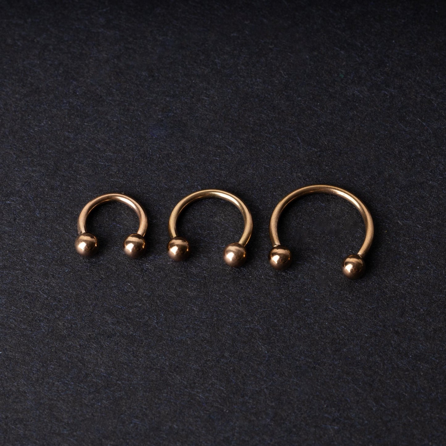 Three rose gold-tone stainless steel horseshoe hoops in varying sizes with screw-on ball ends, displayed against a dark background.