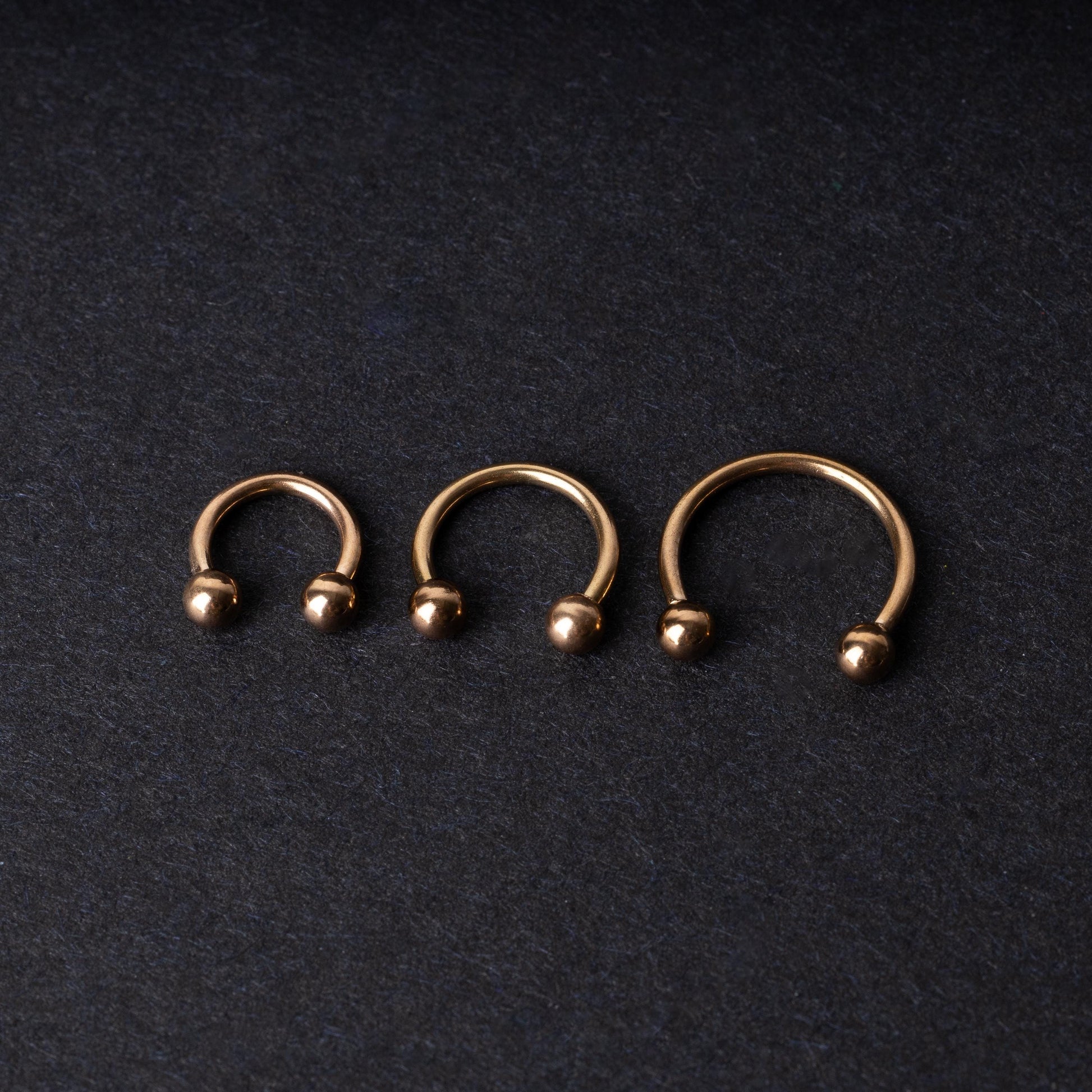 Three rose gold-tone stainless steel horseshoe hoops in varying sizes with screw-on ball ends, displayed against a dark background.