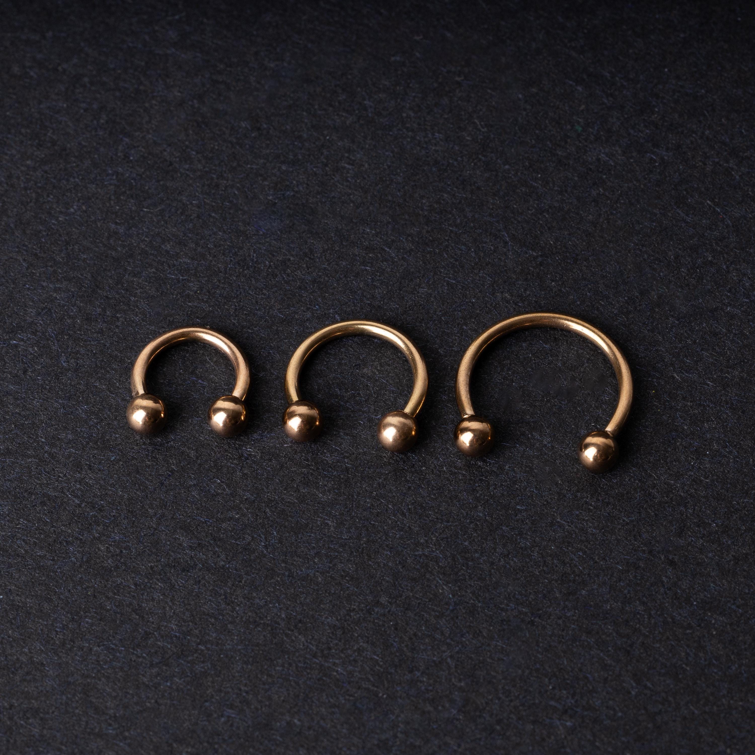 Three rose gold-tone stainless steel horseshoe hoops in varying sizes with screw-on ball ends, displayed against a dark background.