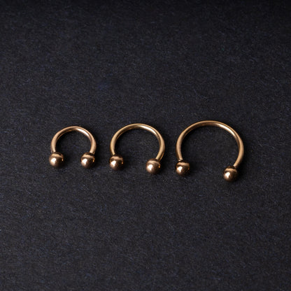 Three rose gold-tone stainless steel horseshoe hoops in varying sizes with screw-on ball ends, displayed against a dark background.