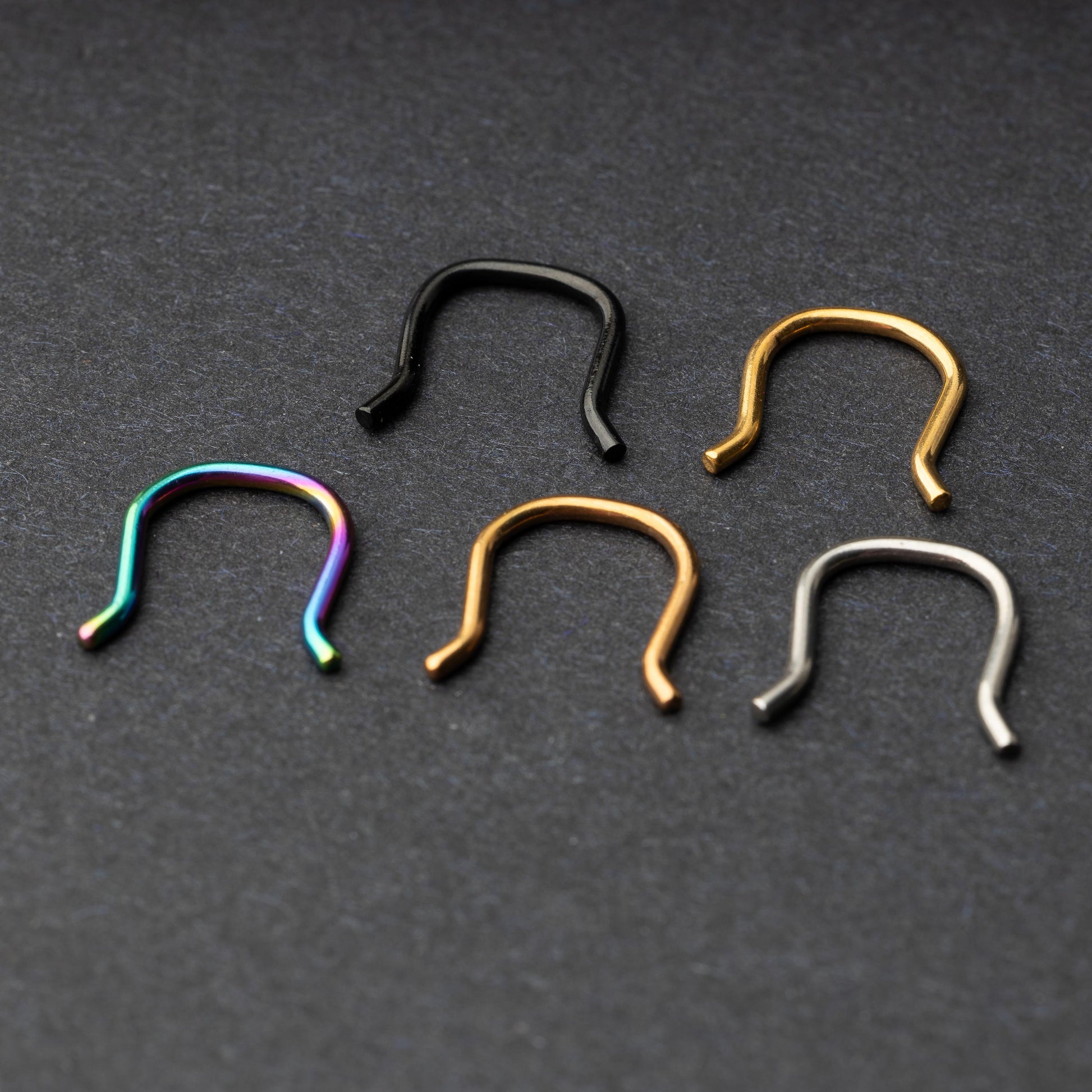 Five U-shaped septum retainers in black, gold, silver, and rainbow finishes, arranged on a dark background.
