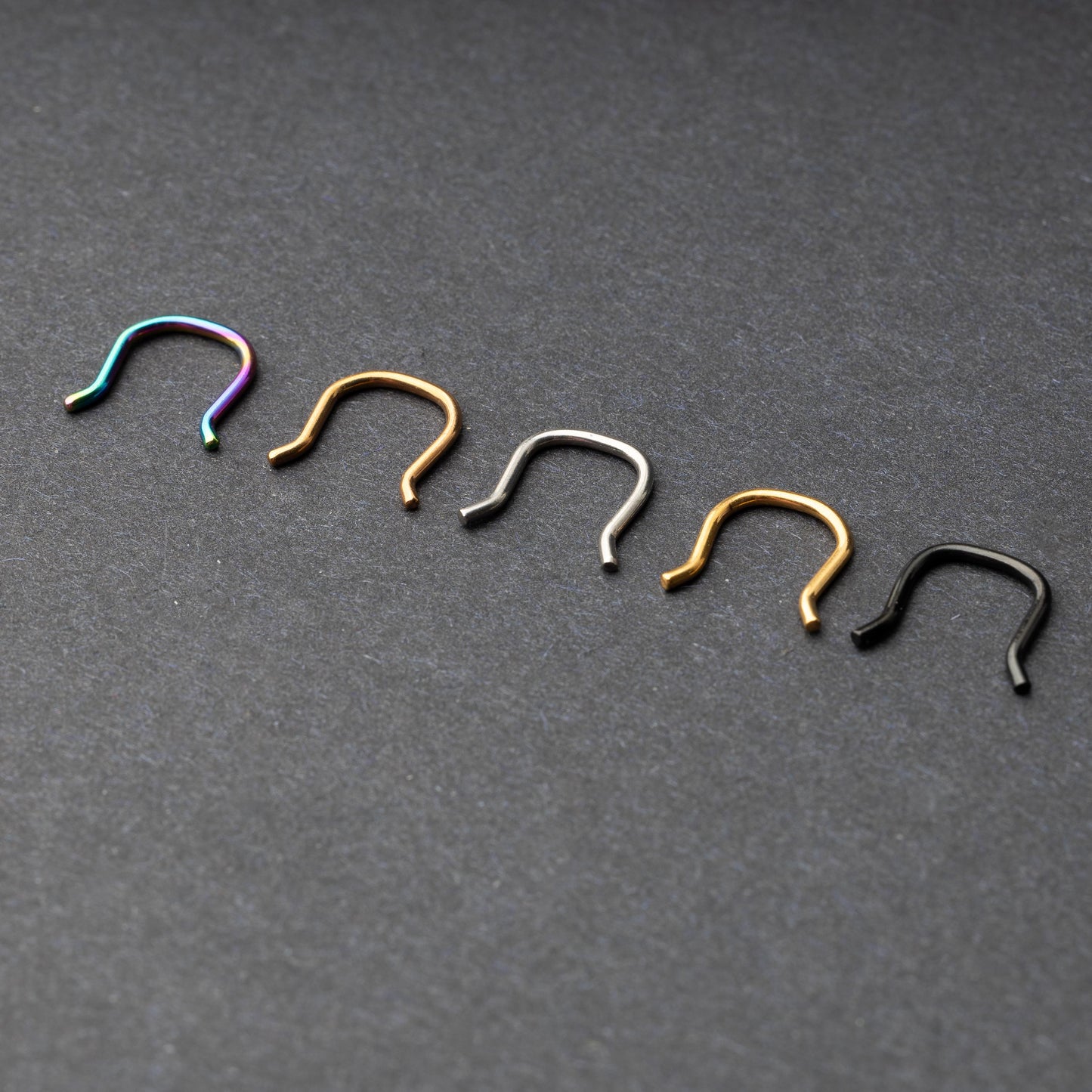 Five U-shaped septum retainers in black, gold, silver, and rainbow finishes, arranged on a dark background.