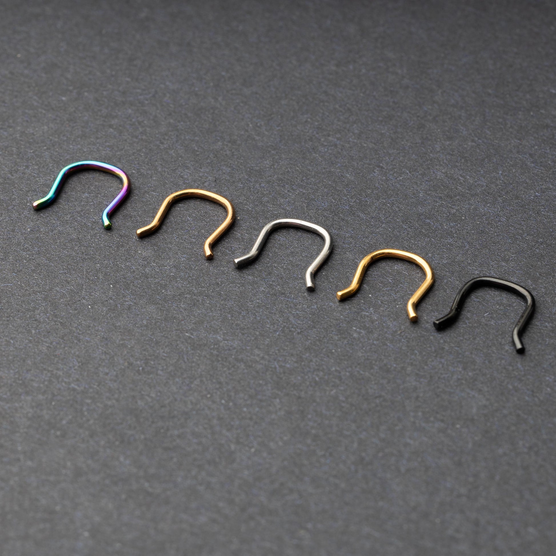 Five U-shaped septum retainers in black, gold, silver, and rainbow finishes, arranged on a dark background.