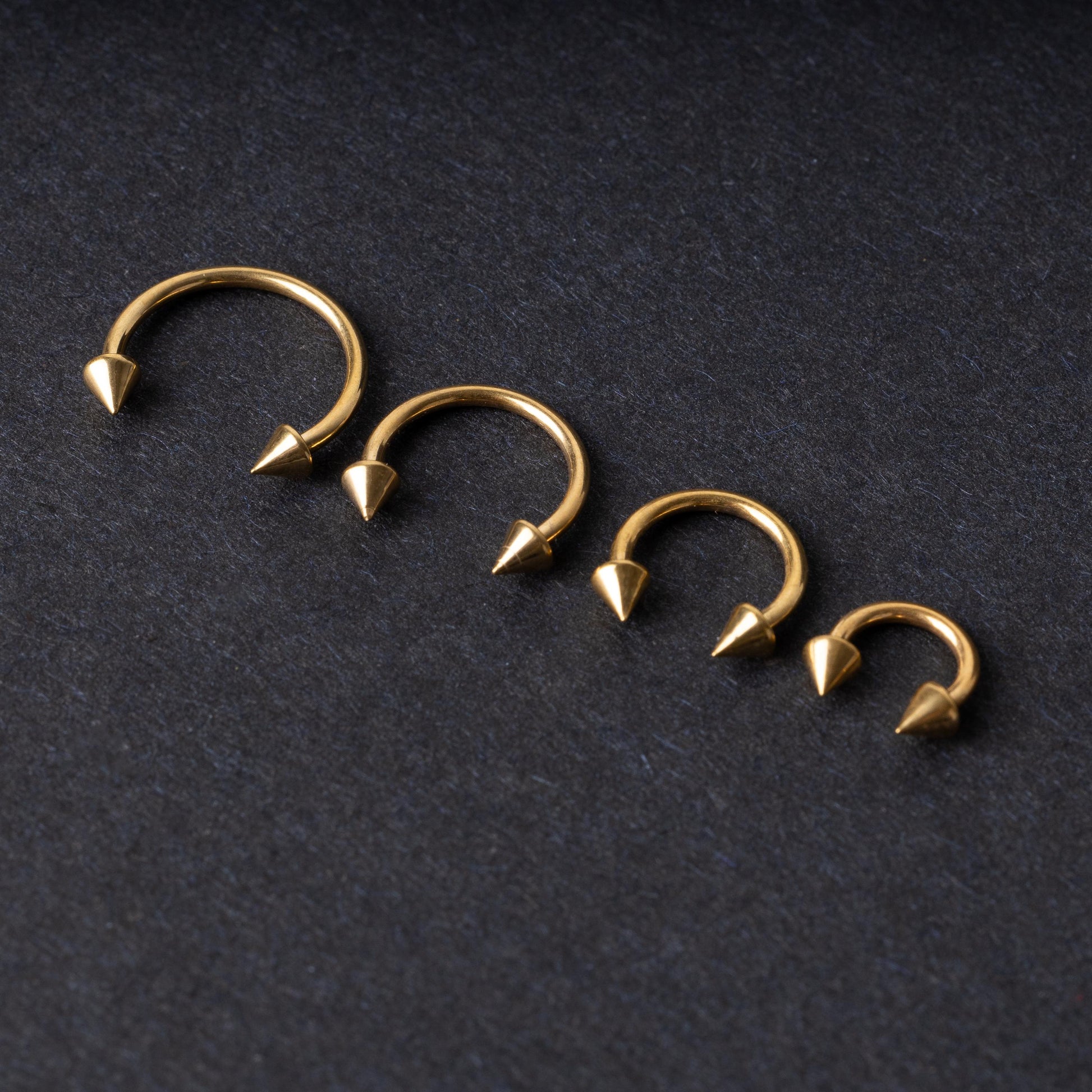 Various sized gold titanium circular barbells with spiked ends displayed on a dark background, arranged by size from large to small.