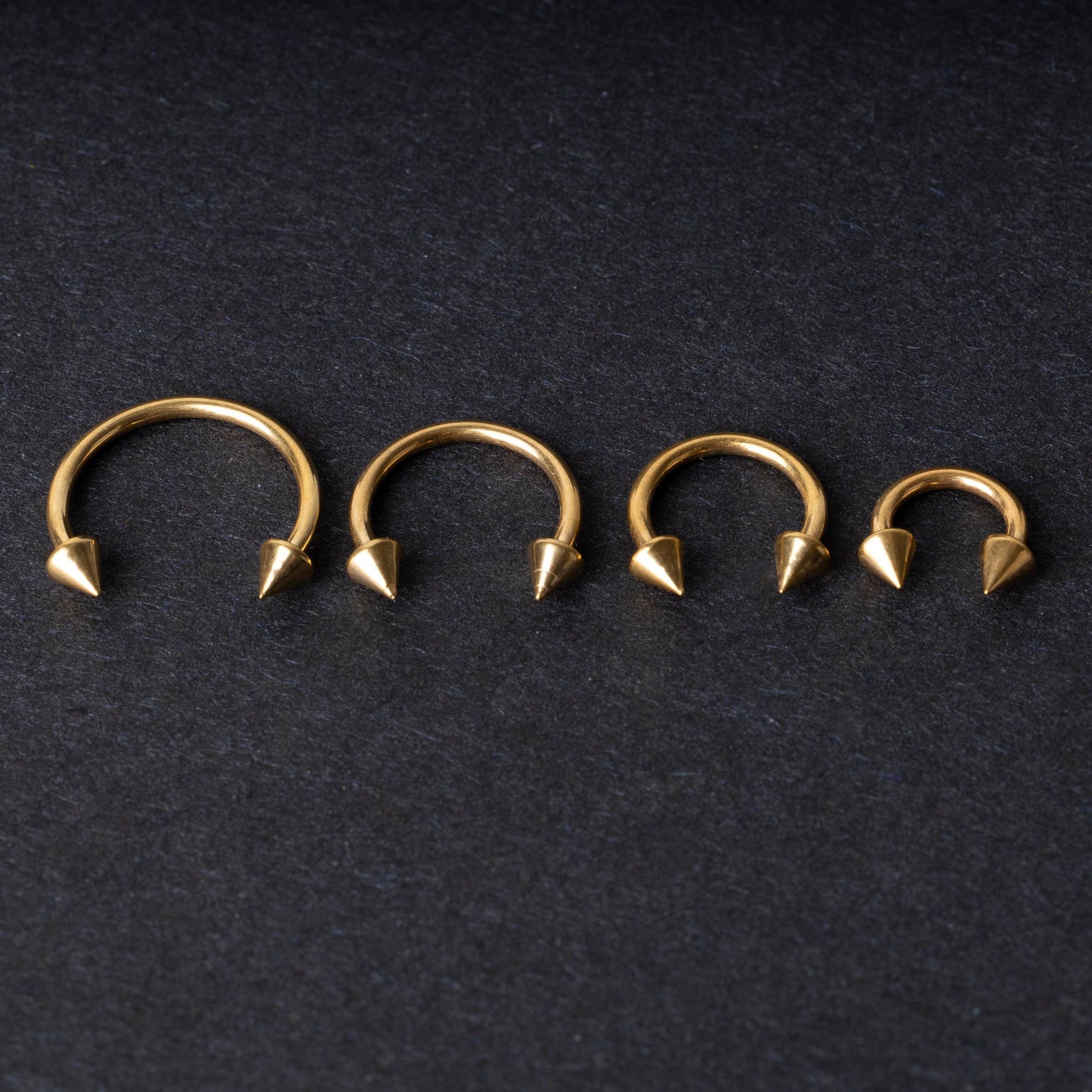 Various sized gold titanium circular barbells with spiked ends displayed on a dark background, arranged by size from large to small.