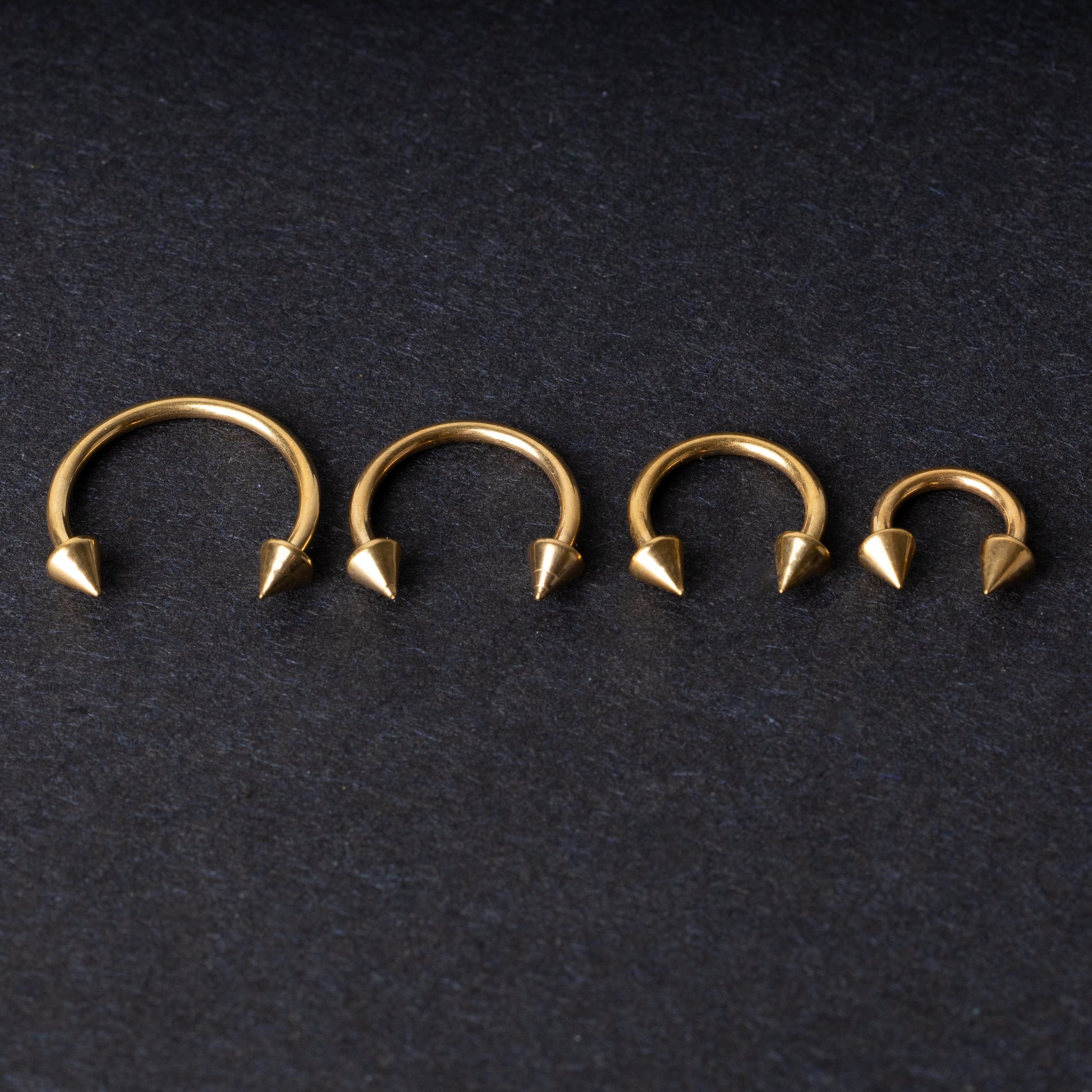 Various sized gold titanium circular barbells with spiked ends displayed on a dark background, arranged by size from large to small.