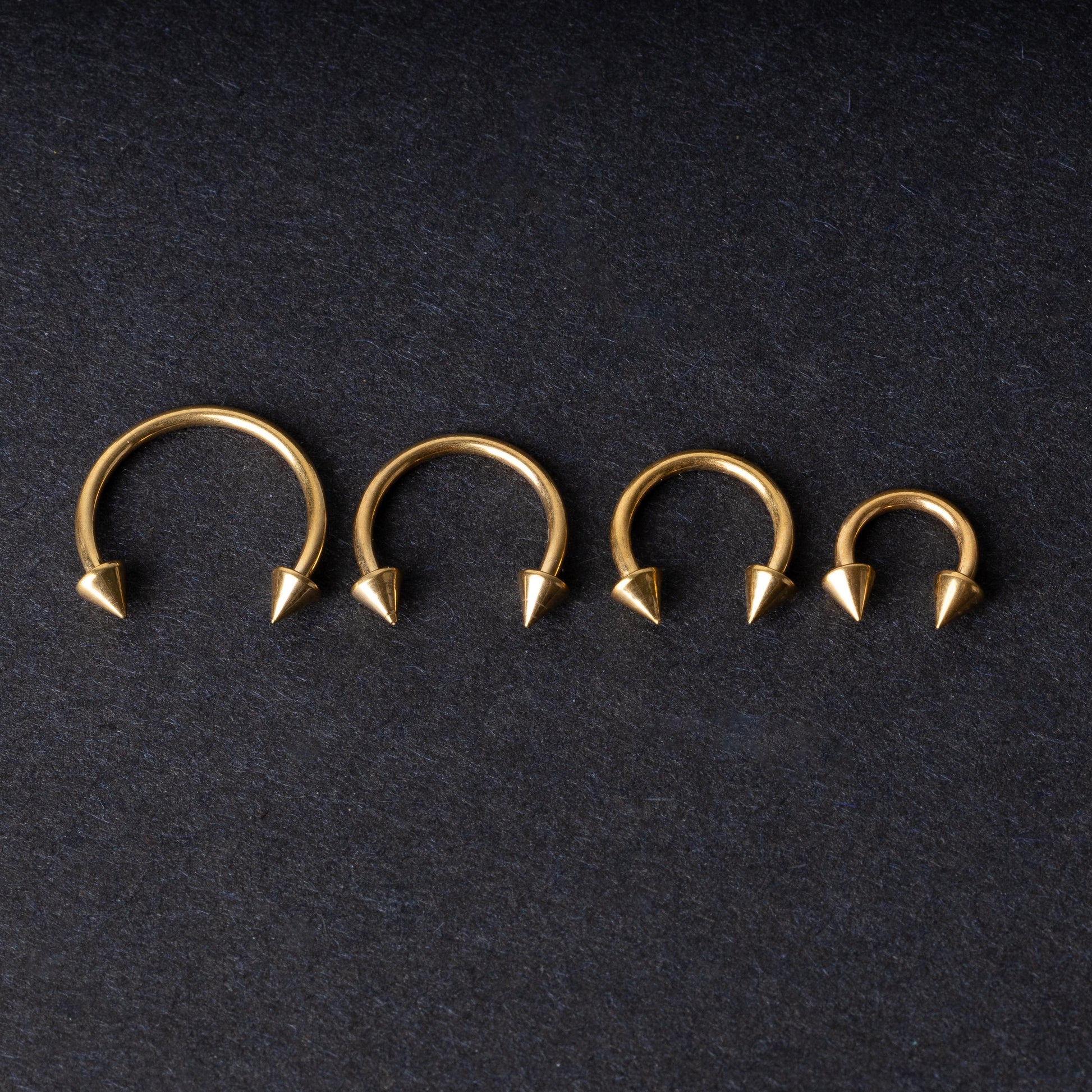 Various sized gold titanium circular barbells with spiked ends displayed on a dark background, arranged by size from large to small.