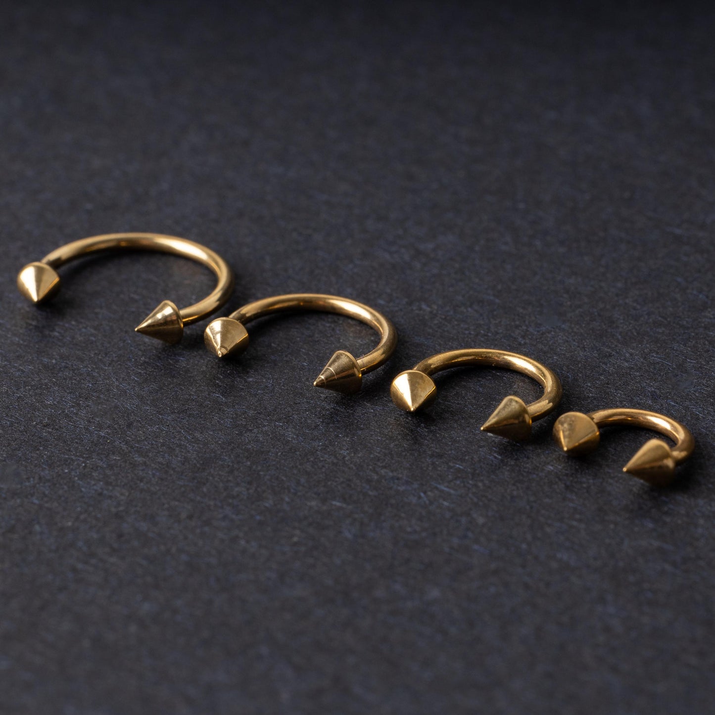 Various sized gold titanium circular barbells with spiked ends displayed on a dark background, arranged by size from large to small.