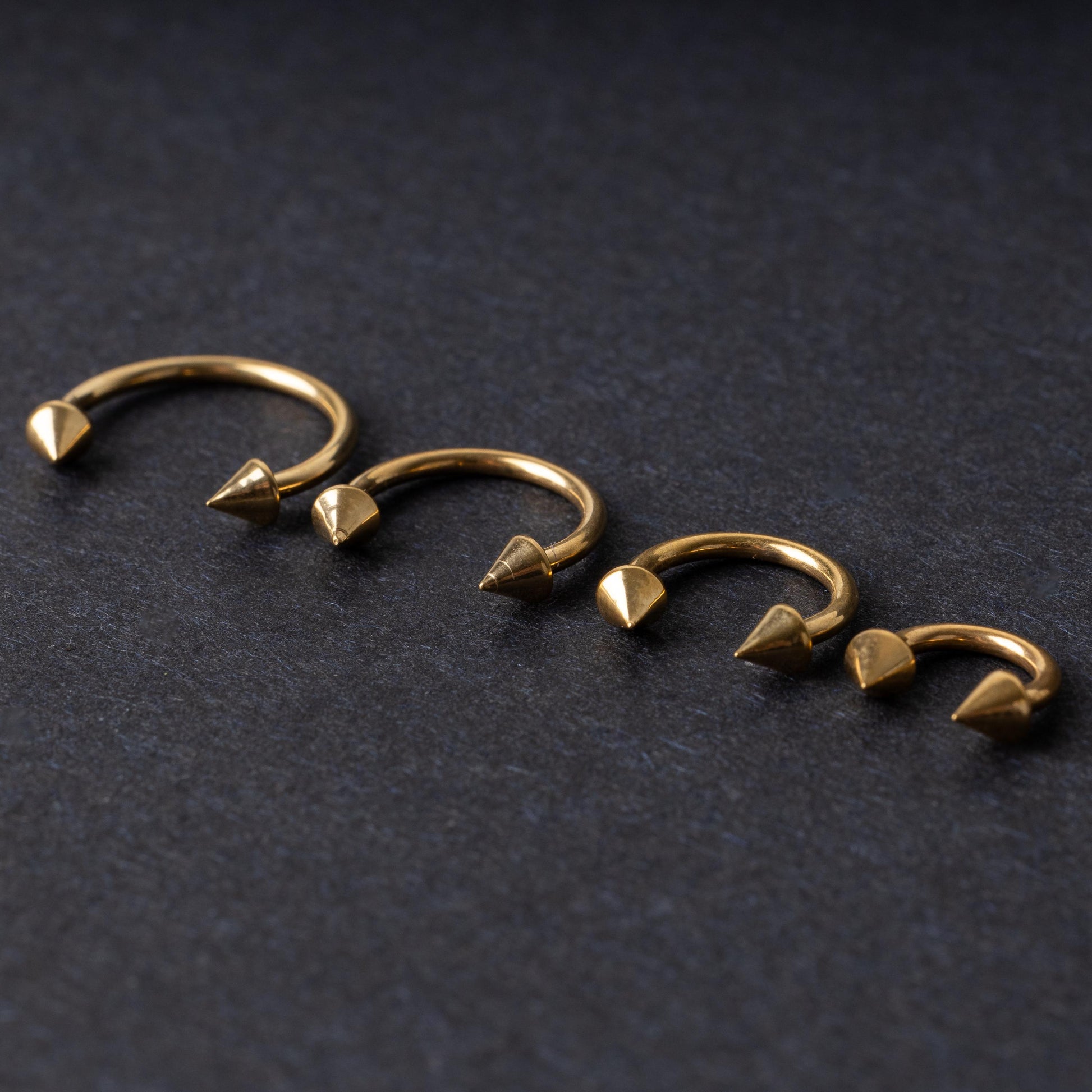Various sized gold titanium circular barbells with spiked ends displayed on a dark background, arranged by size from large to small.