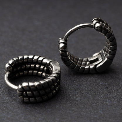 Two black metal hoop earrings with a bold, textured grid design, displayed on a dark background.