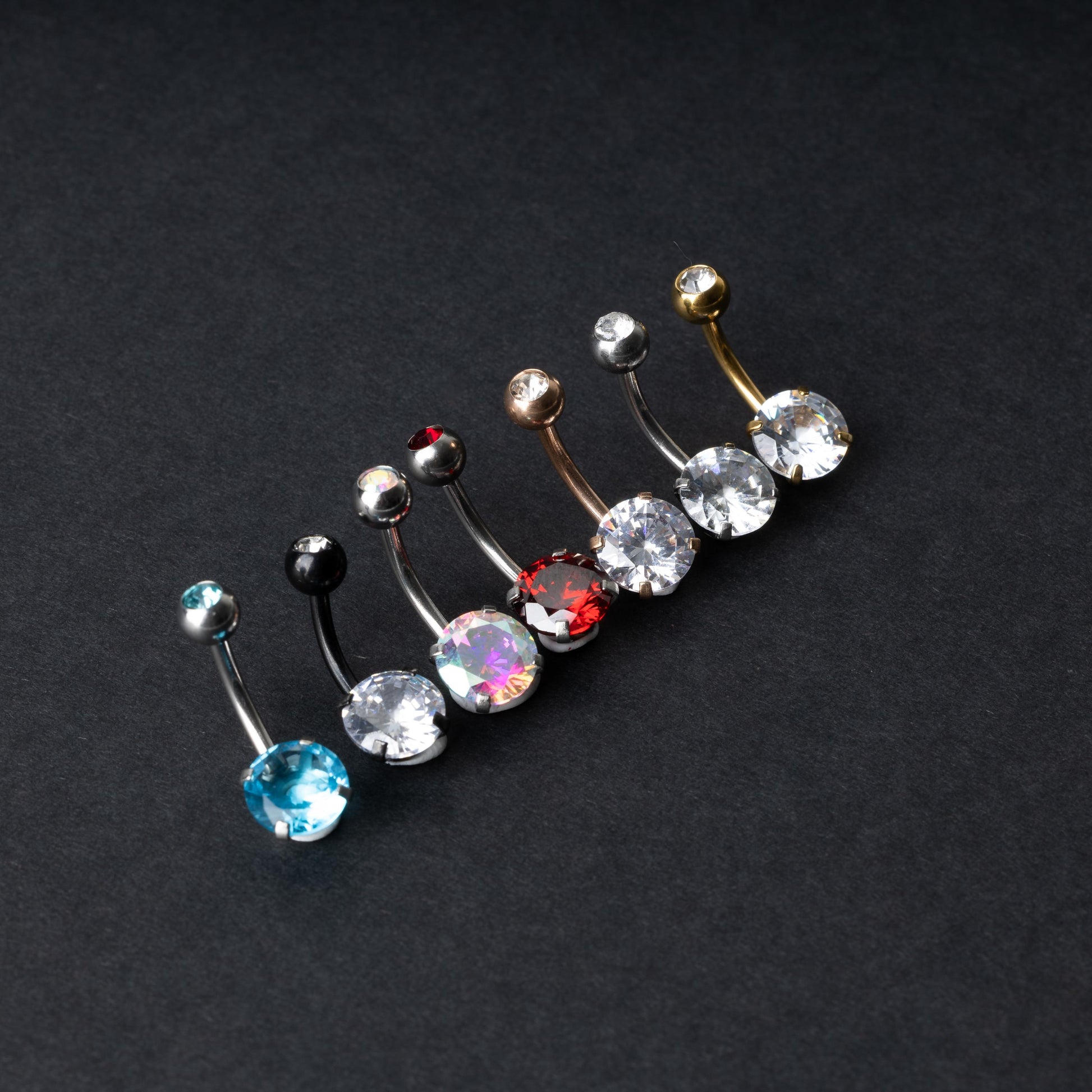 A collection of colorful belly button rings with curved barbells, featuring large gemstone centerpieces in shades of blue, black, clear, iridescent, red, and gold, displayed on a black background.