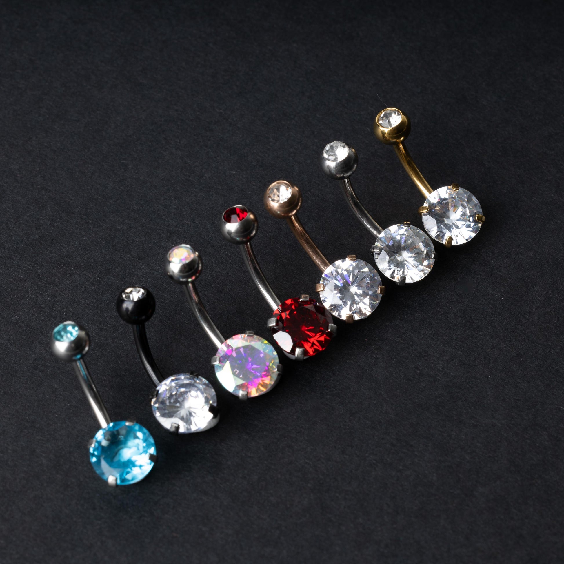 A collection of colorful belly button rings with curved barbells, featuring large gemstone centerpieces in shades of blue, black, clear, iridescent, red, and gold, displayed on a black background.