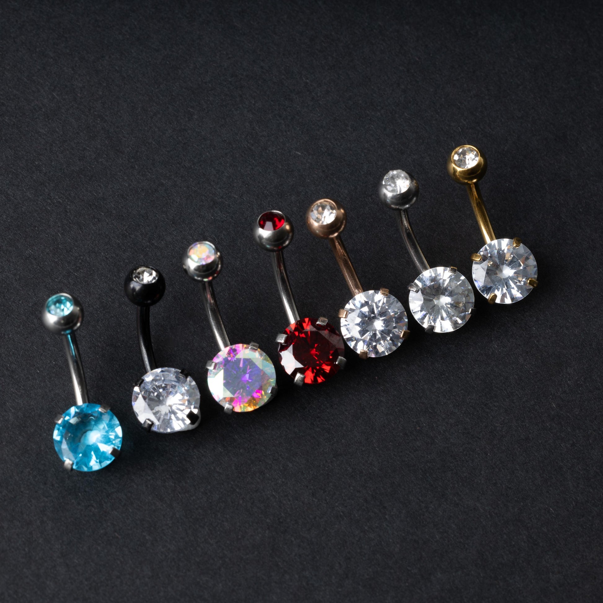 A collection of colorful belly button rings with curved barbells, featuring large gemstone centerpieces in shades of blue, black, clear, iridescent, red, and gold, displayed on a black background.