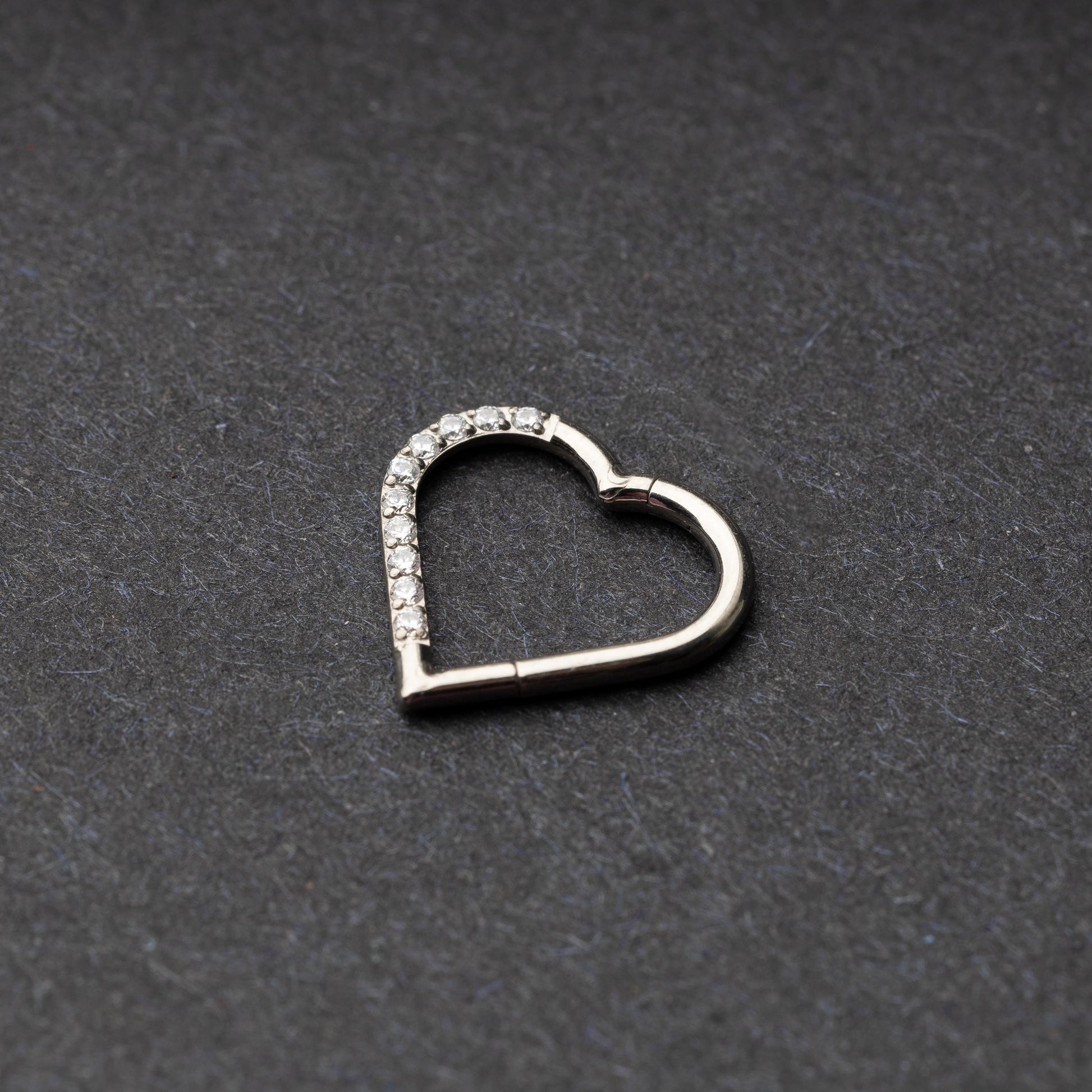 Heart-shaped daith clicker with crystal accents along one side, displayed on a dark textured background.
