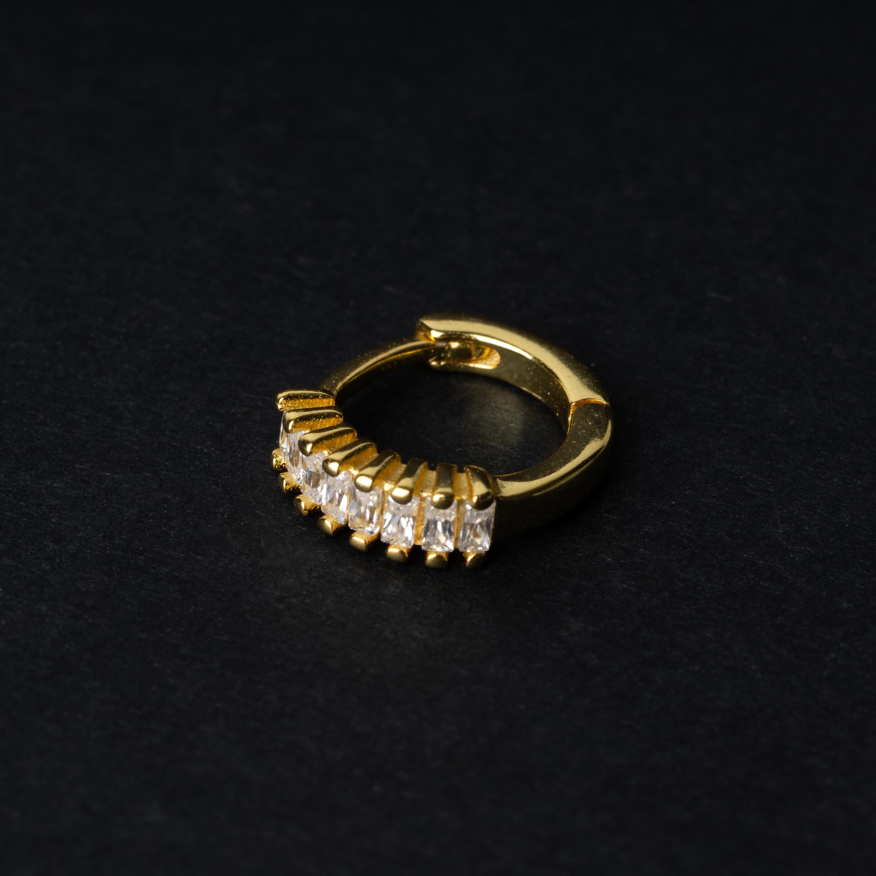 A gold septum clicker featuring a row of sparkling cubic zirconia gemstones, displayed on a black background.
