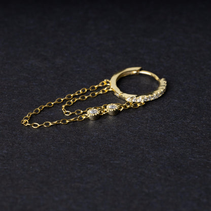 A gold septum clicker with a decorative design featuring dual chains and cubic zirconia gems, displayed on a black background.