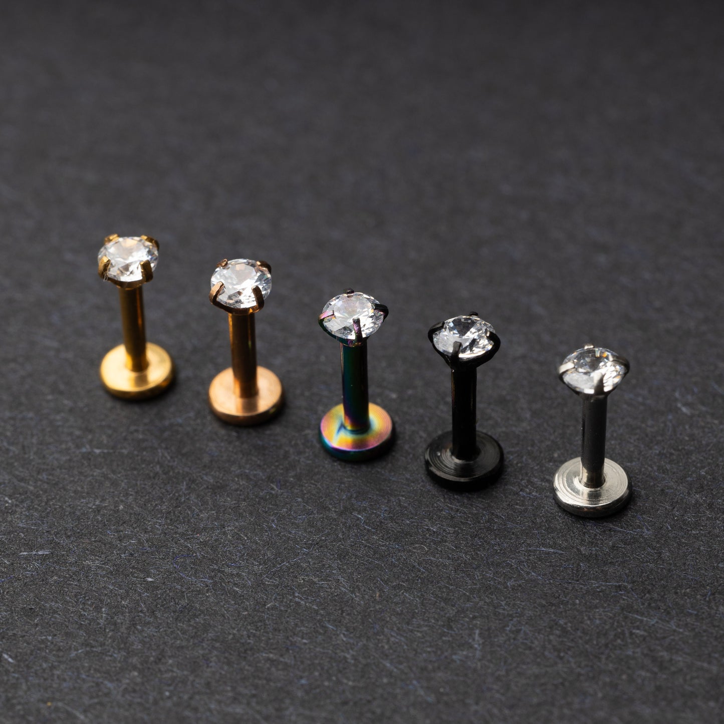 A set of five labret studs with clear gemstones, featuring finishes in gold, rose gold, rainbow, black, and silver, displayed on a dark background.