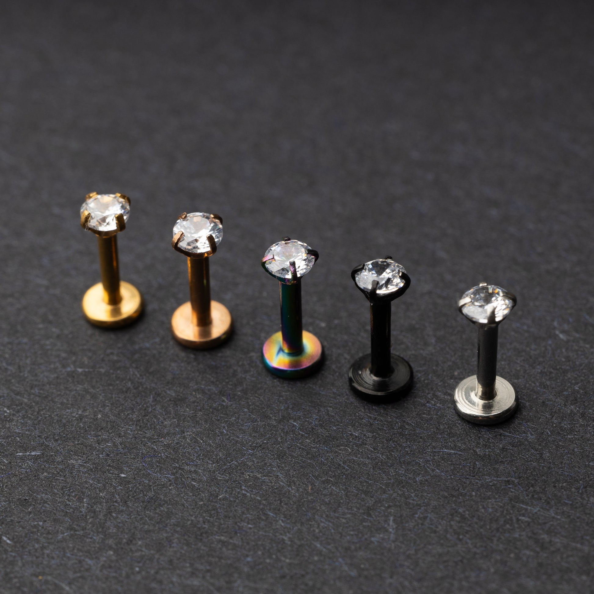 A set of five labret studs with clear gemstones, featuring finishes in gold, rose gold, rainbow, black, and silver, displayed on a dark background.