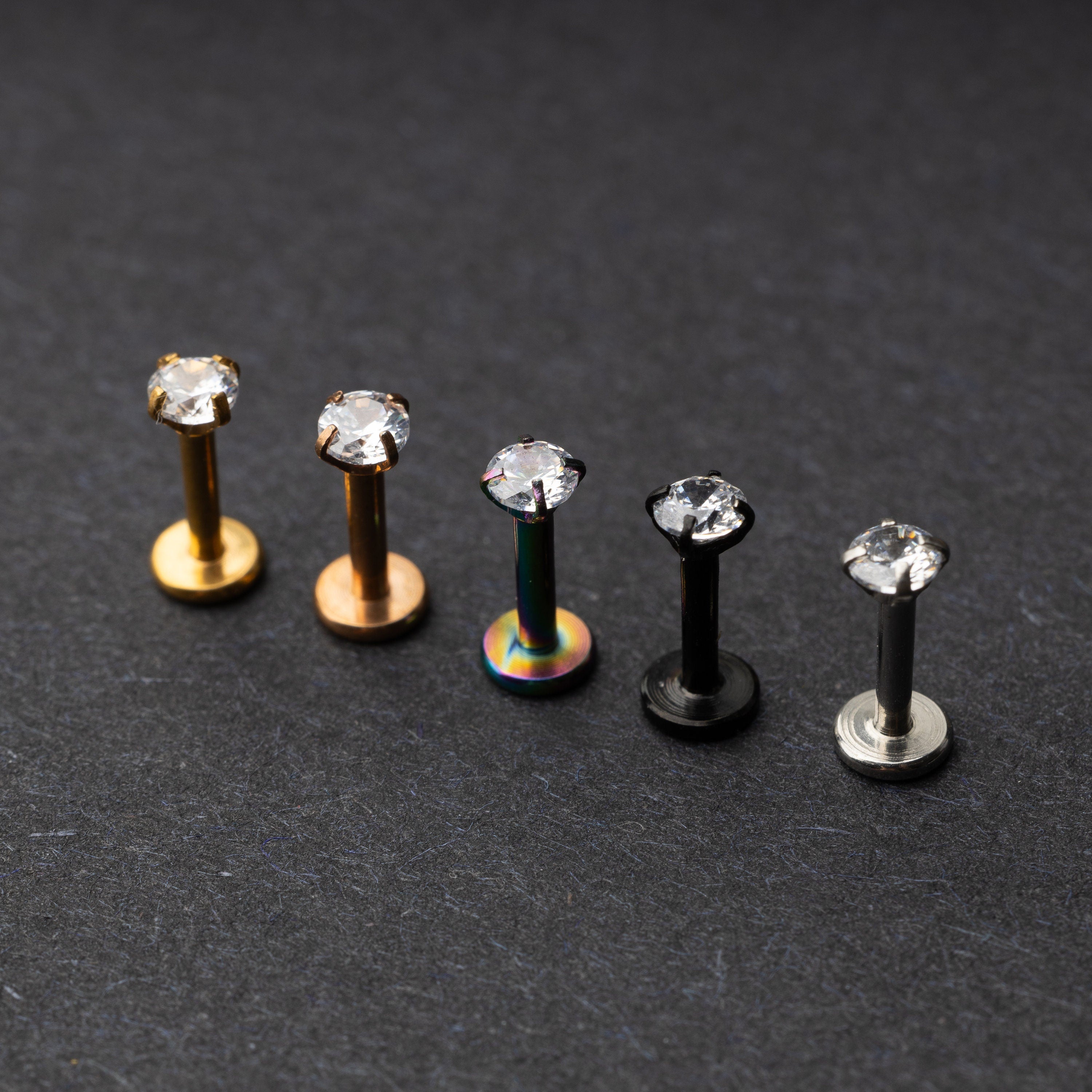 A set of five labret studs with clear gemstones, featuring finishes in gold, rose gold, rainbow, black, and silver, displayed on a dark background.