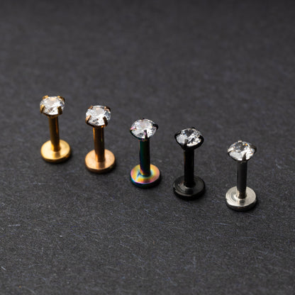 A set of five labret studs with clear gemstones, featuring finishes in gold, rose gold, rainbow, black, and silver, displayed on a dark background.