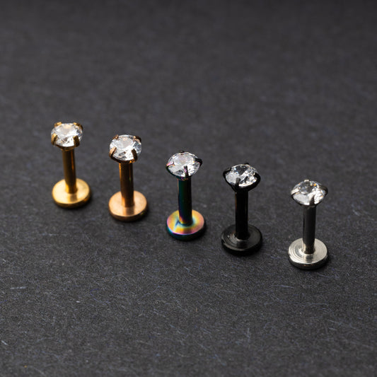 A set of five labret studs with clear gemstones, featuring finishes in gold, rose gold, rainbow, black, and silver, displayed on a dark background.