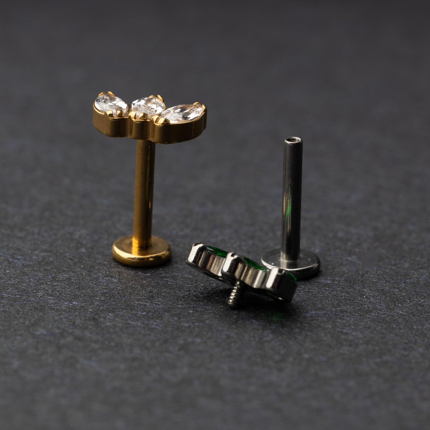 Two flat back bars featuring a triple gem design: one with emerald green gems on a silver base and the other with clear gems on a gold base, displayed on a dark background.
