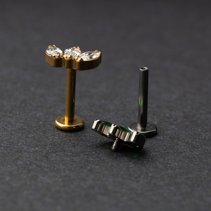 Two flat back bars featuring a triple gem design: one with emerald green gems on a silver base and the other with clear gems on a gold base, displayed on a dark background.
