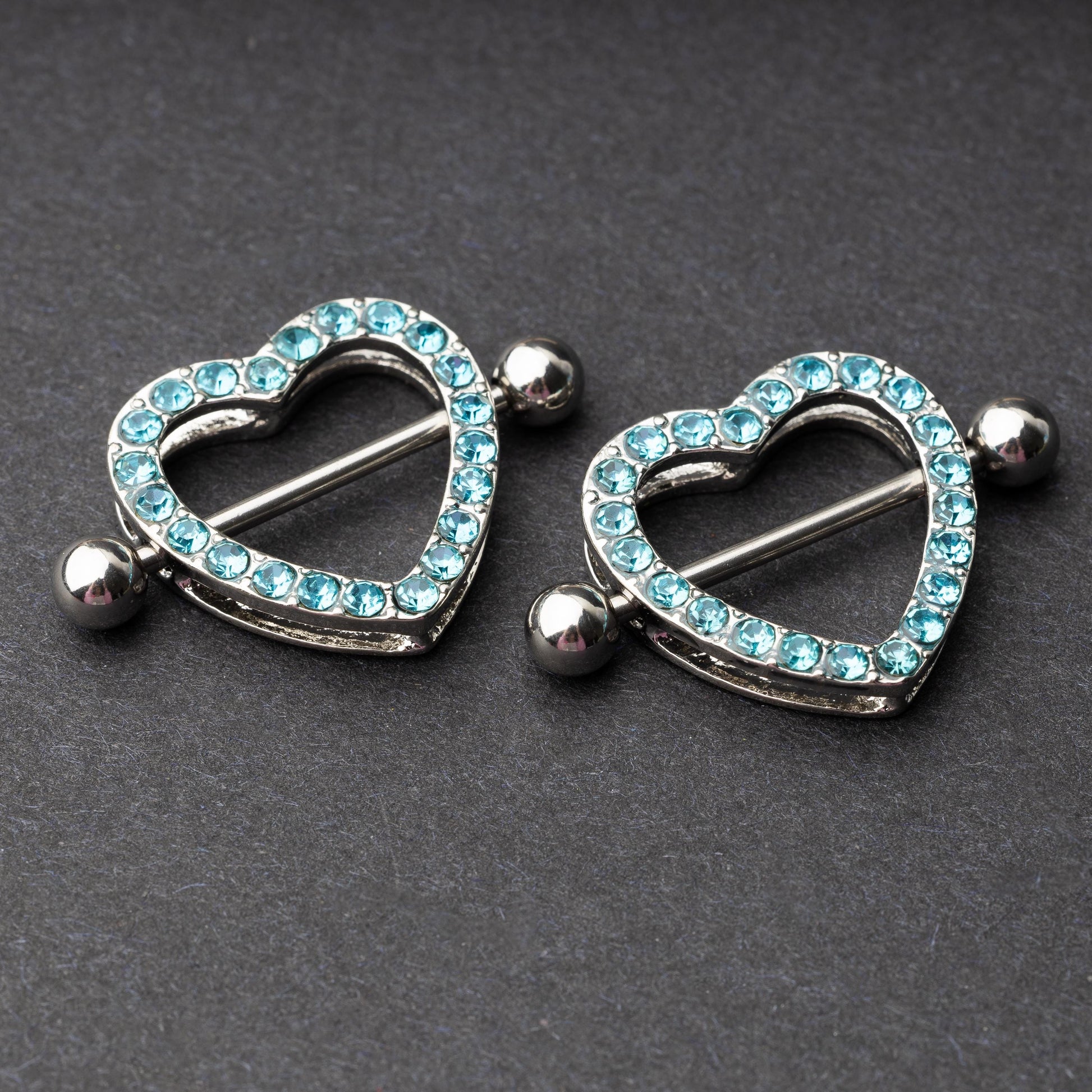 Two heart-shaped nipple barbells with silver metal and light blue gemstone accents, displayed on a dark background.