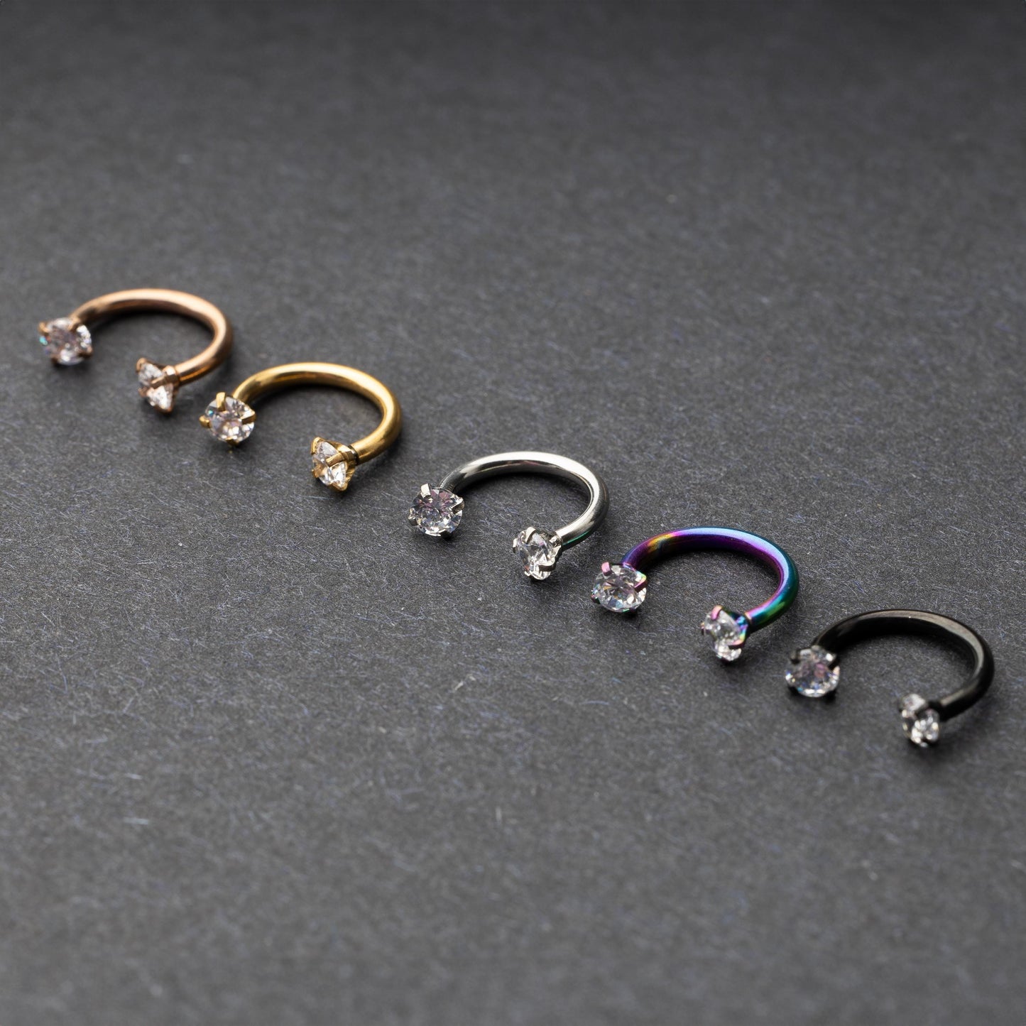 A set of five horseshoe barbells in finishes including rose gold, black, gold, rainbow, and silver, each with gemstone ends, displayed on a dark background.