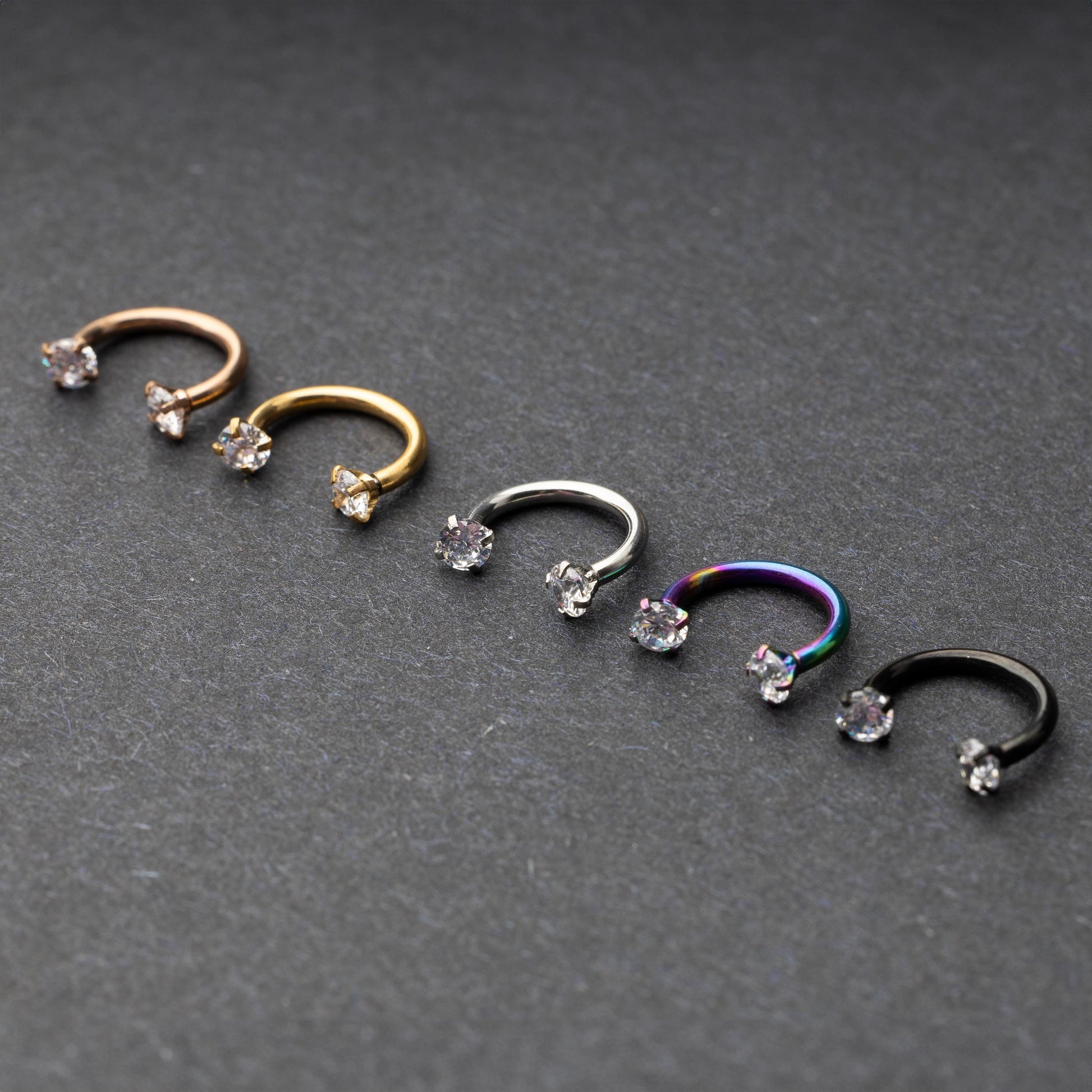 A set of five horseshoe barbells in finishes including rose gold, black, gold, rainbow, and silver, each with gemstone ends, displayed on a dark background.