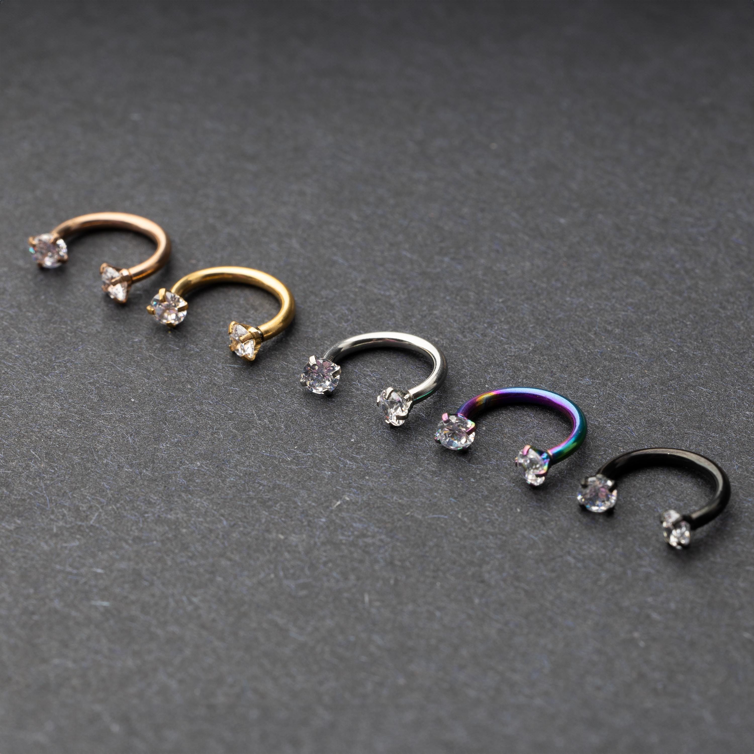A set of five horseshoe barbells in finishes including rose gold, black, gold, rainbow, and silver, each with gemstone ends, displayed on a dark background.
