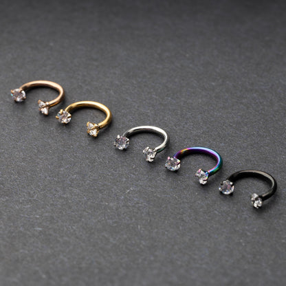 A set of five horseshoe barbells in finishes including rose gold, black, gold, rainbow, and silver, each with gemstone ends, displayed on a dark background.