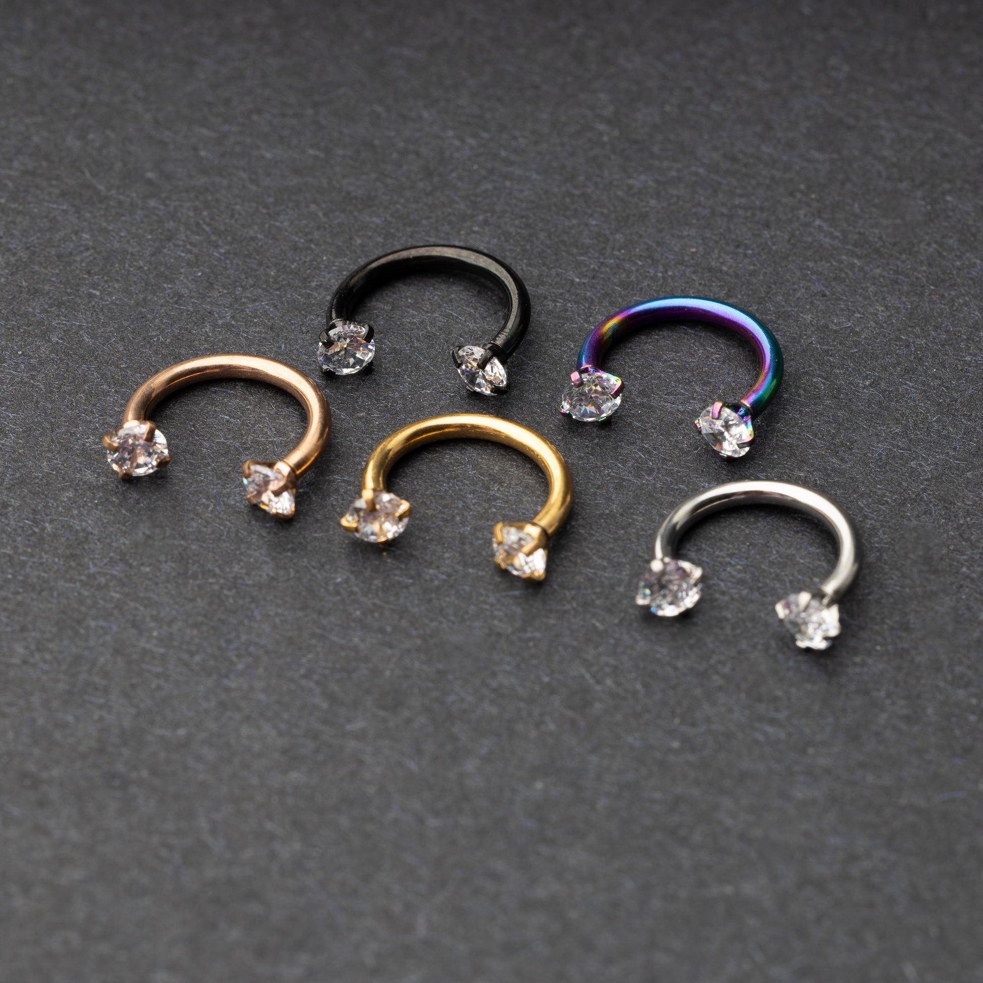 A set of five horseshoe barbells in finishes including rose gold, black, gold, rainbow, and silver, each with gemstone ends, displayed on a dark background.
