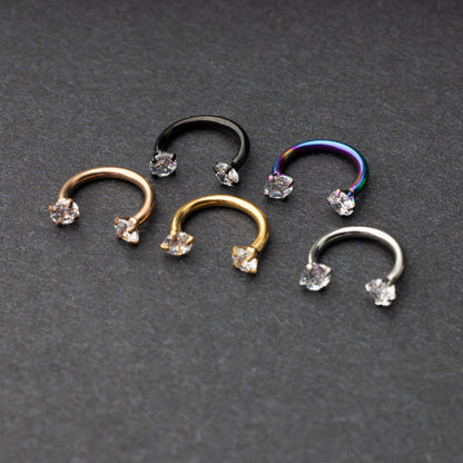 A set of five horseshoe barbells in finishes including rose gold, black, gold, rainbow, and silver, each with gemstone ends, displayed on a dark background.
