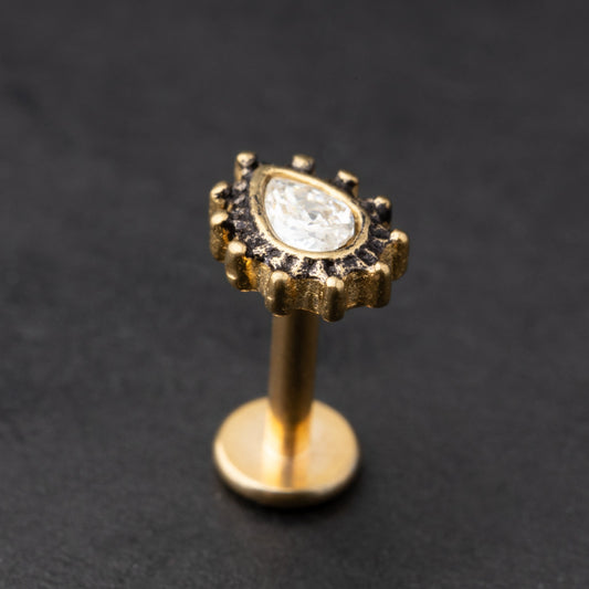 Gold labret stud with a teardrop-shaped crystal centerpiece and detailed border, displayed on a dark background.