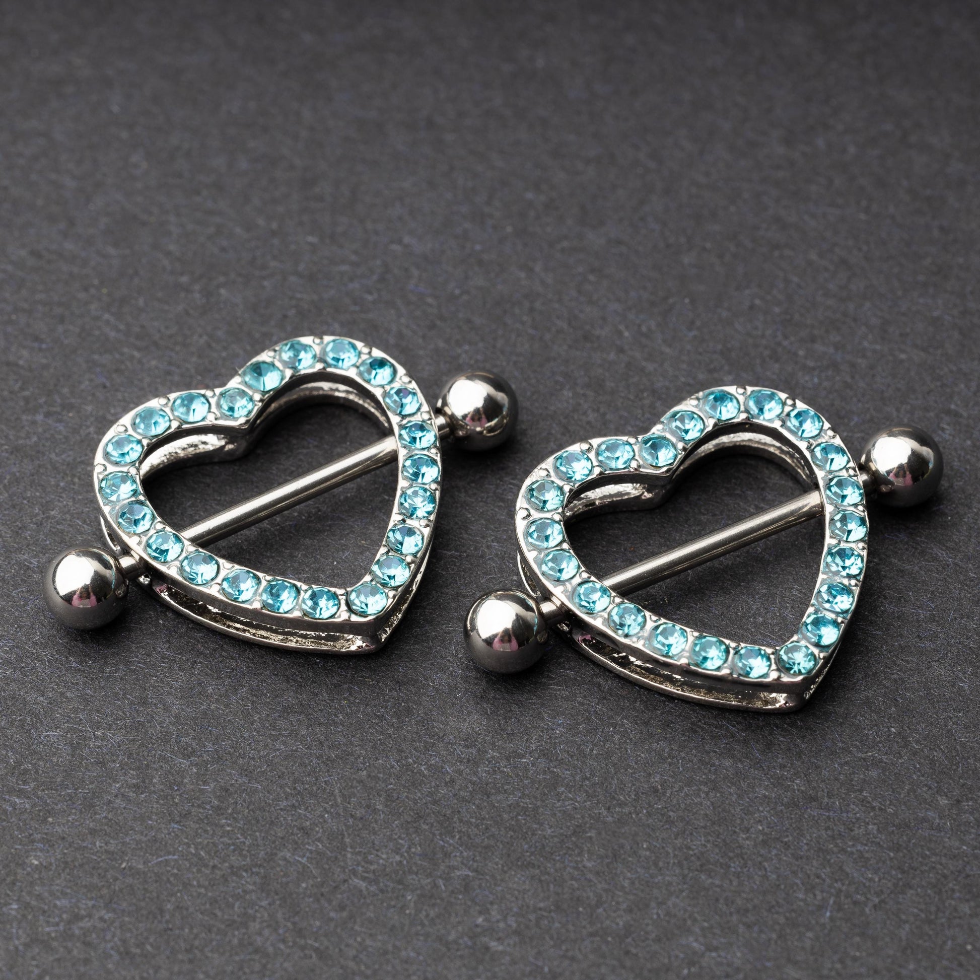 Two heart-shaped nipple barbells with silver metal and light blue gemstone accents, displayed on a dark background.