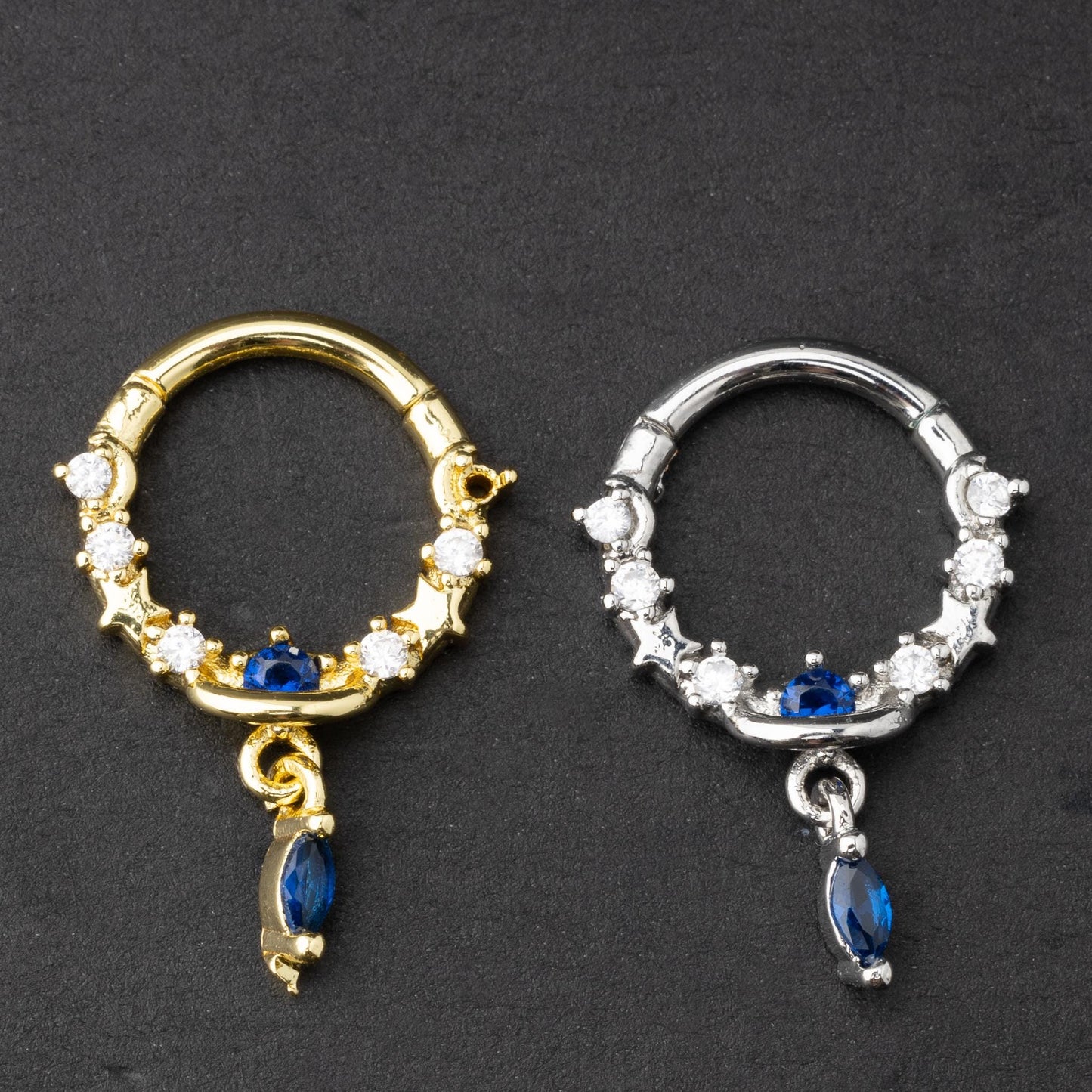 Gold and silver septum clickers adorned with cubic zirconia and a dangling blue gemstone charm, displayed on a dark background.