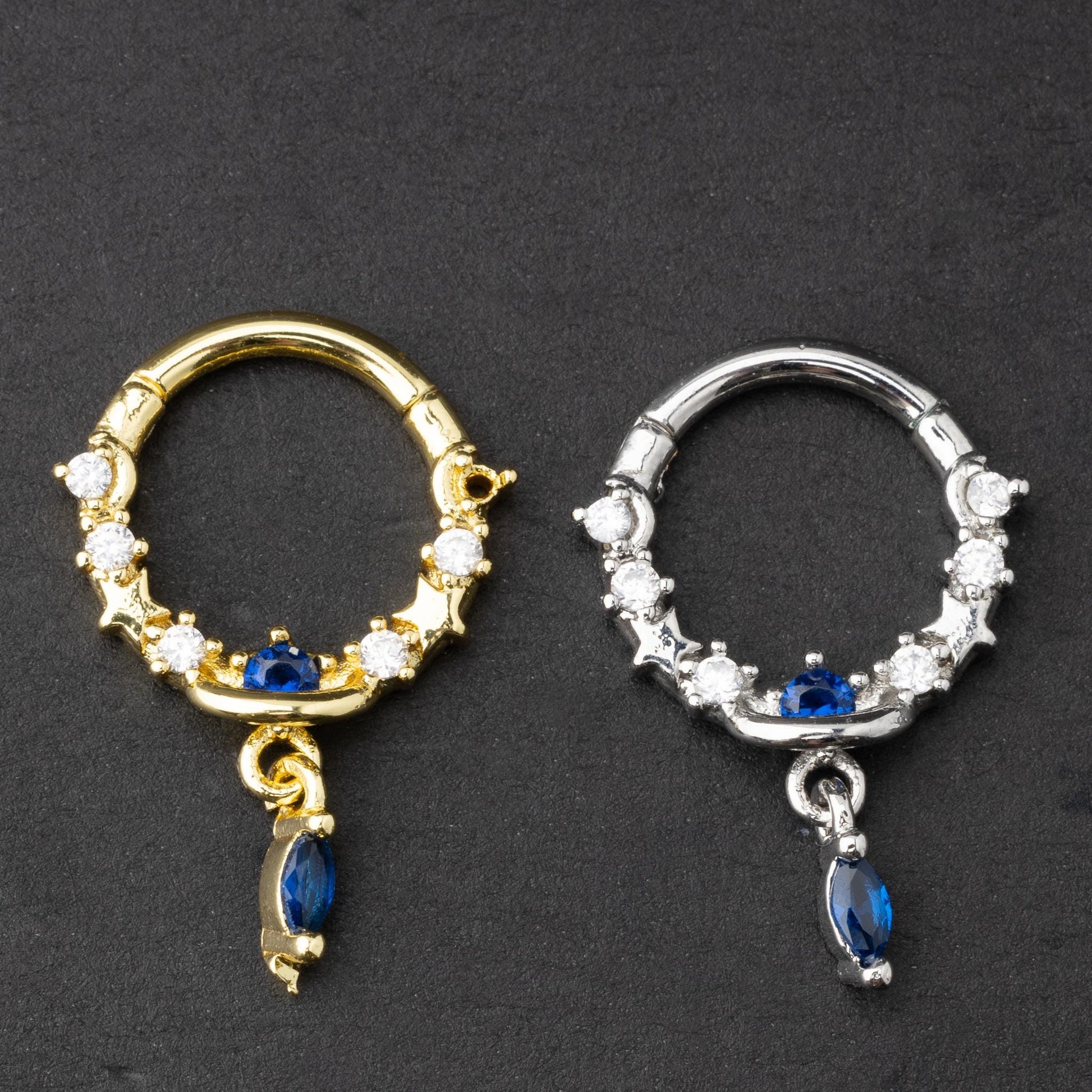 Gold and silver septum clickers adorned with cubic zirconia and a dangling blue gemstone charm, displayed on a dark background.