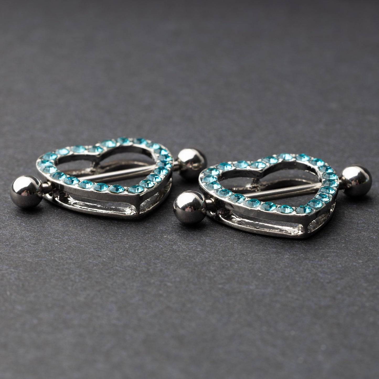 Two heart-shaped nipple barbells with silver metal and light blue gemstone accents, displayed on a dark background.