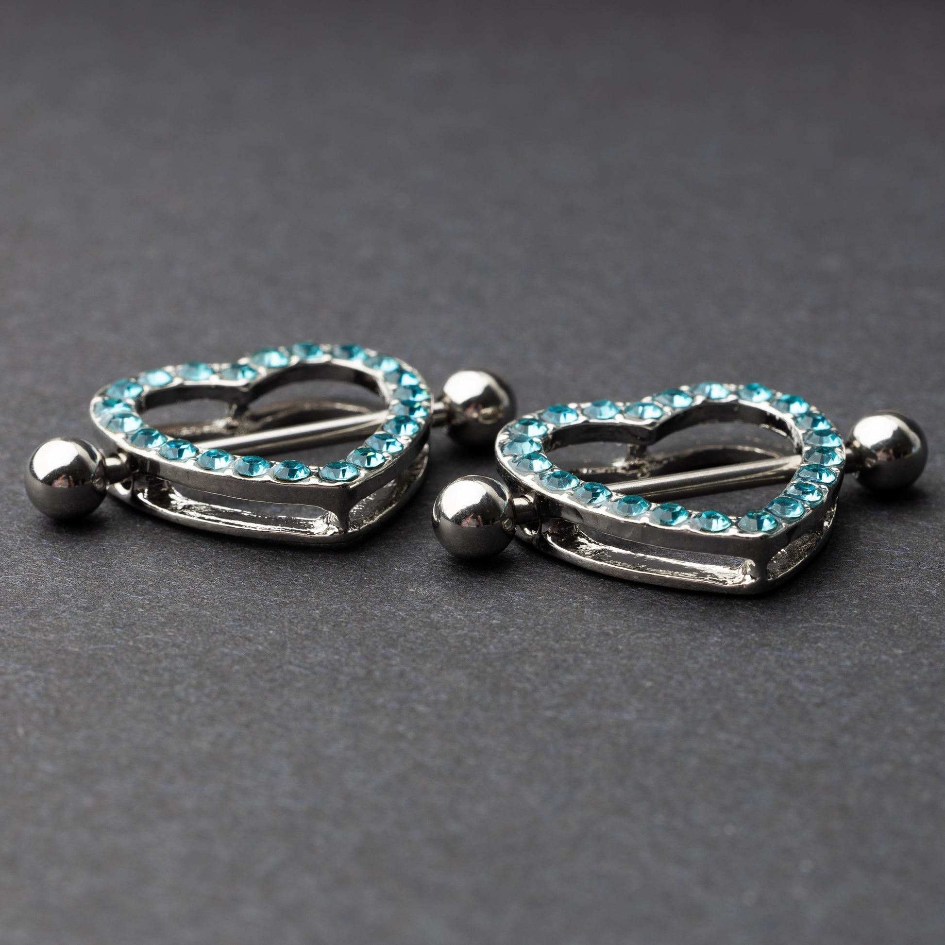 Two heart-shaped nipple barbells with silver metal and light blue gemstone accents, displayed on a dark background.