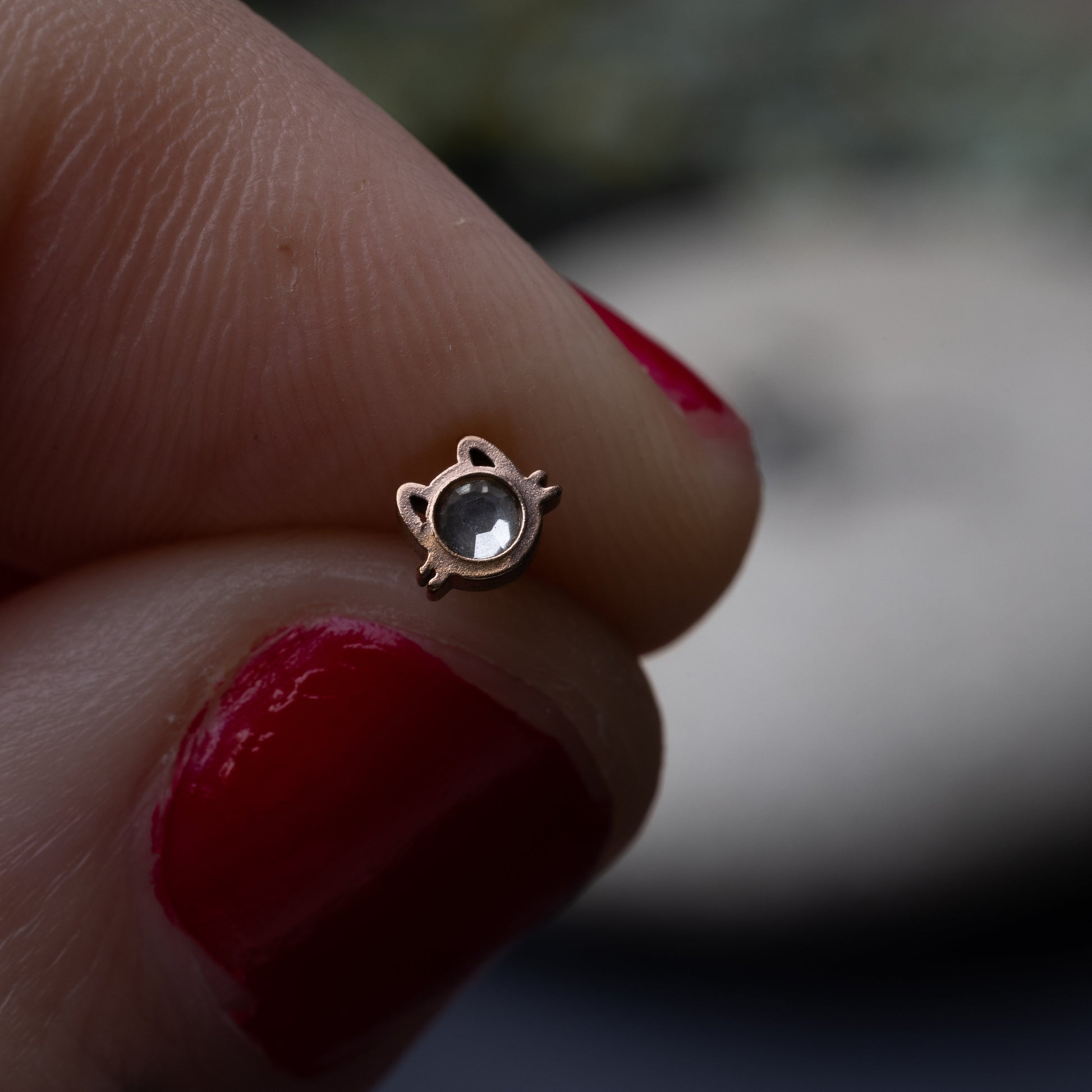 A rose gold stud earring in the shape of a cat’s face with a bezel-set gemstone, held between a thinger and thumb. 