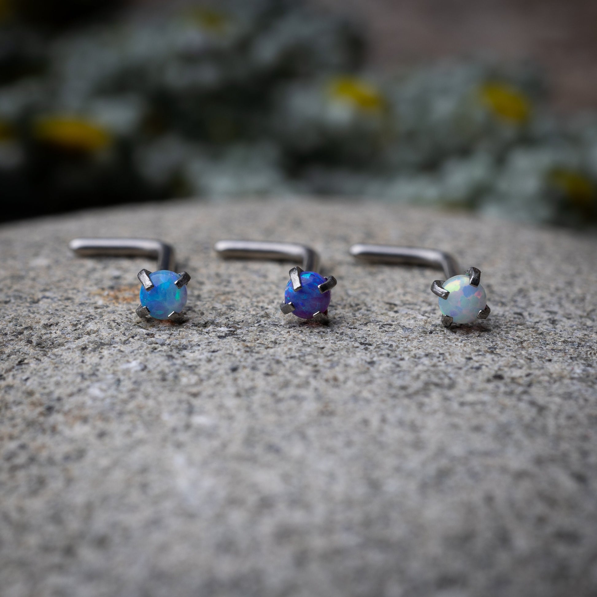 Three L-shaped opalescent nose studs in different shades, displayed on a stone background.